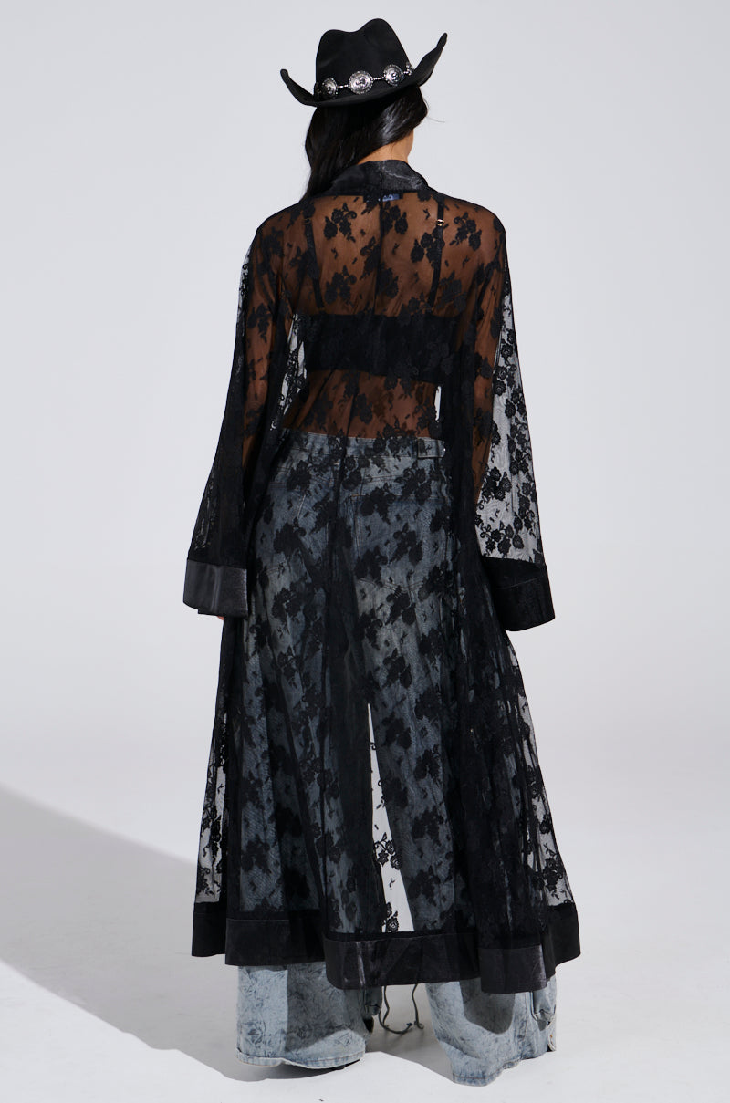 Wearing the Bora Bora Lace Duster in black sheer floral lace over light blue jeans, a person stands with their back to the camera. They complete the festival-ready look with a decorated black cowboy hat against a plain gray background.