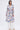 A woman stands against a plain white background wearing the PLANT THE SEED SHEER TRENCH, a long, floral print sheer trench coat with a tie belt, styled over wide-legged white pants and open-toed sandals.