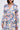 Wearing the PLANT THE SEED SHEER TRENCH, an organza coat with a pink, blue, and yellow floral print, she accents her look with a chunky gold chain necklace while her shoulder-length dark hair frames her face.