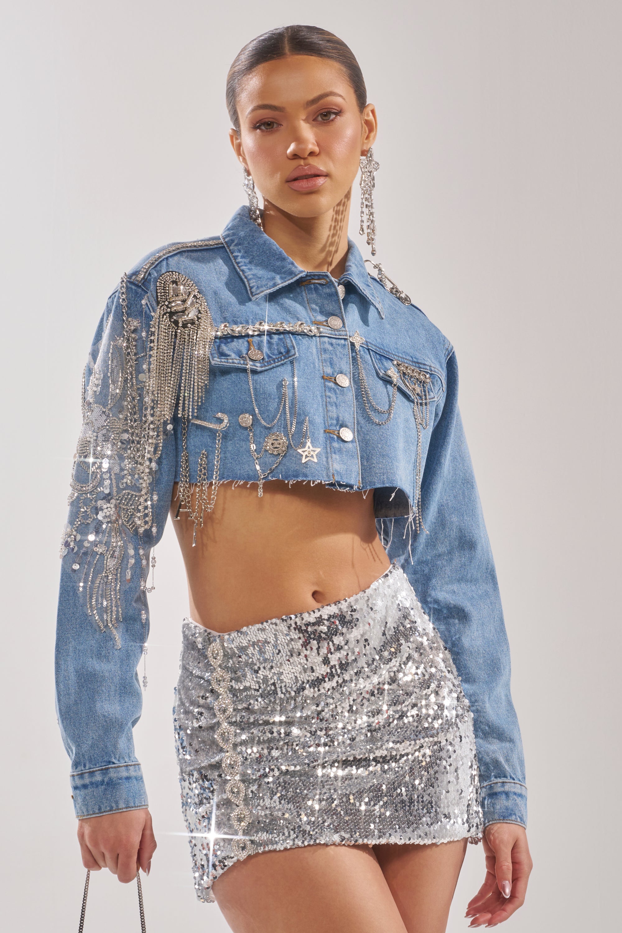A woman models the PIPER EMBELLISHED DENIM JACKET, featuring silver chains and jewels, styled with a shiny silver sequin mini skirt, statement earrings, and a small handbag against a neutral background. 0