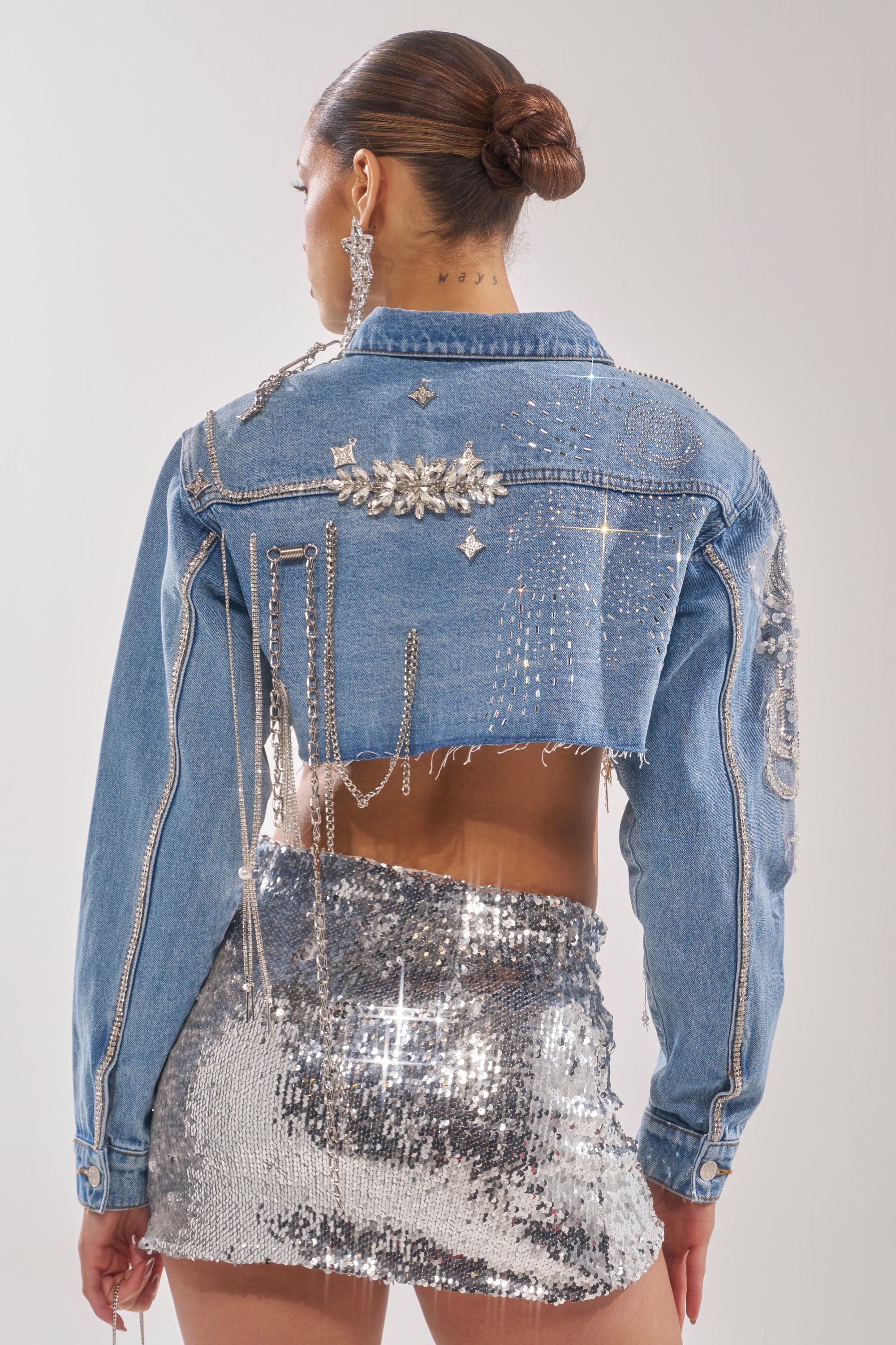 Seen from behind, a woman with her hair in a bun wears the PIPER EMBELLISHED DENIM JACKET, covered in rhinestones and chains, paired with a shiny silver sequin mini skirt and dangling earrings. 9