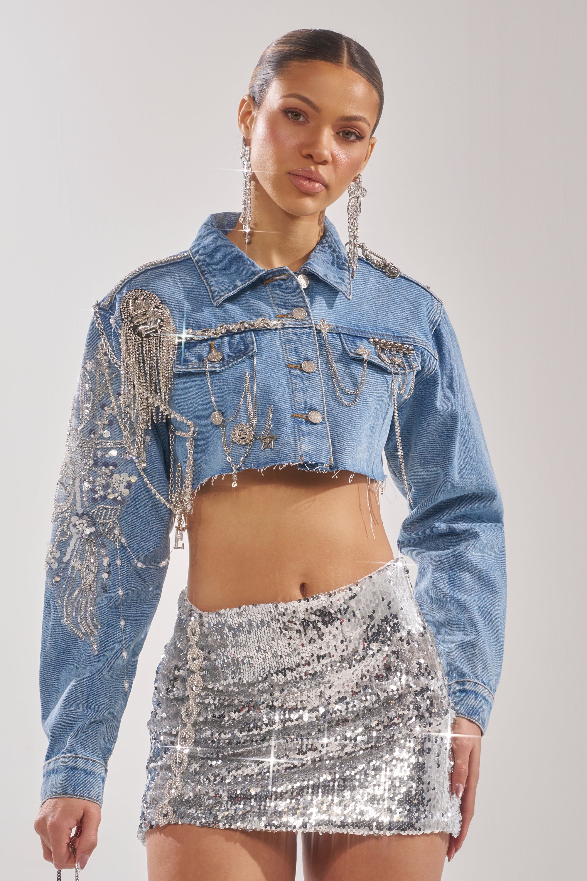 A woman wears the PIPER EMBELLISHED DENIM JACKET, featuring sparkling details and fringe, with a silver sequin mini skirt. She accessorizes with long earrings and has her hair tied back. 3