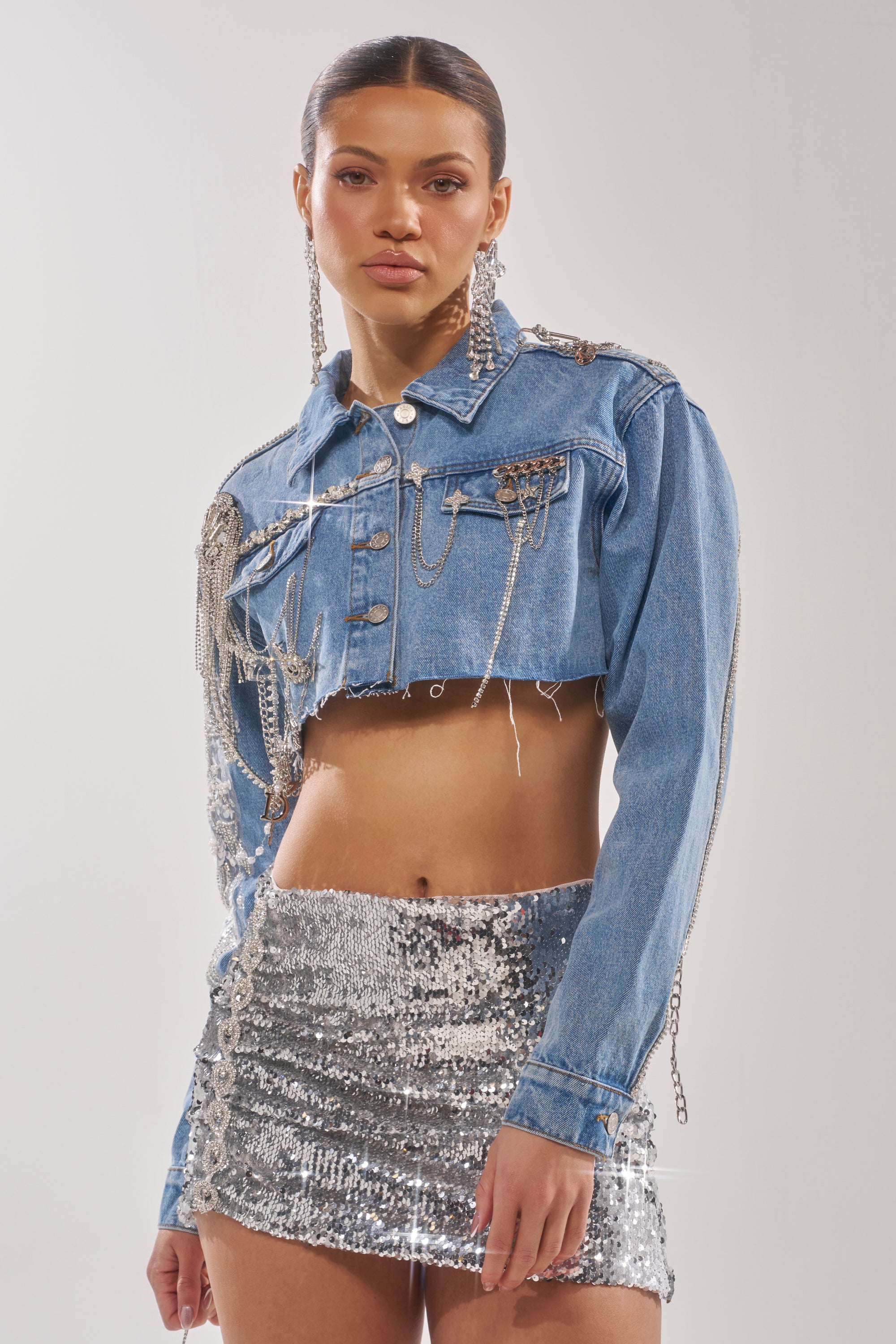 A woman stands against a plain light background wearing the PIPER EMBELLISHED DENIM JACKET adorned with chains and paired with a shiny silver sequin mini skirt. 4