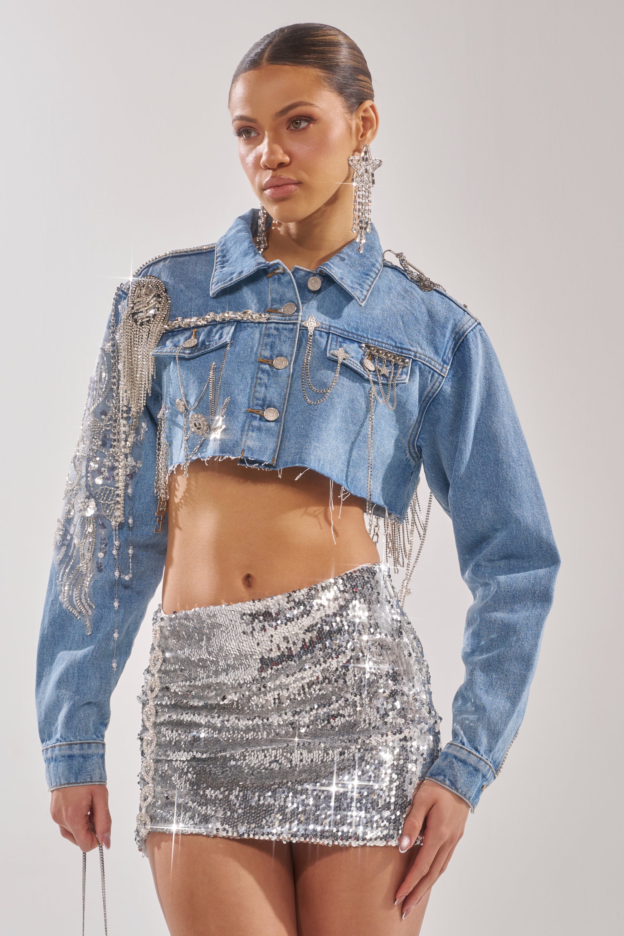 A woman wears the PIPER EMBELLISHED DENIM JACKET, a cropped denim piece with silver fringe and embellishments, styled with a shiny silver sequin mini skirt and sparkly earrings. 5