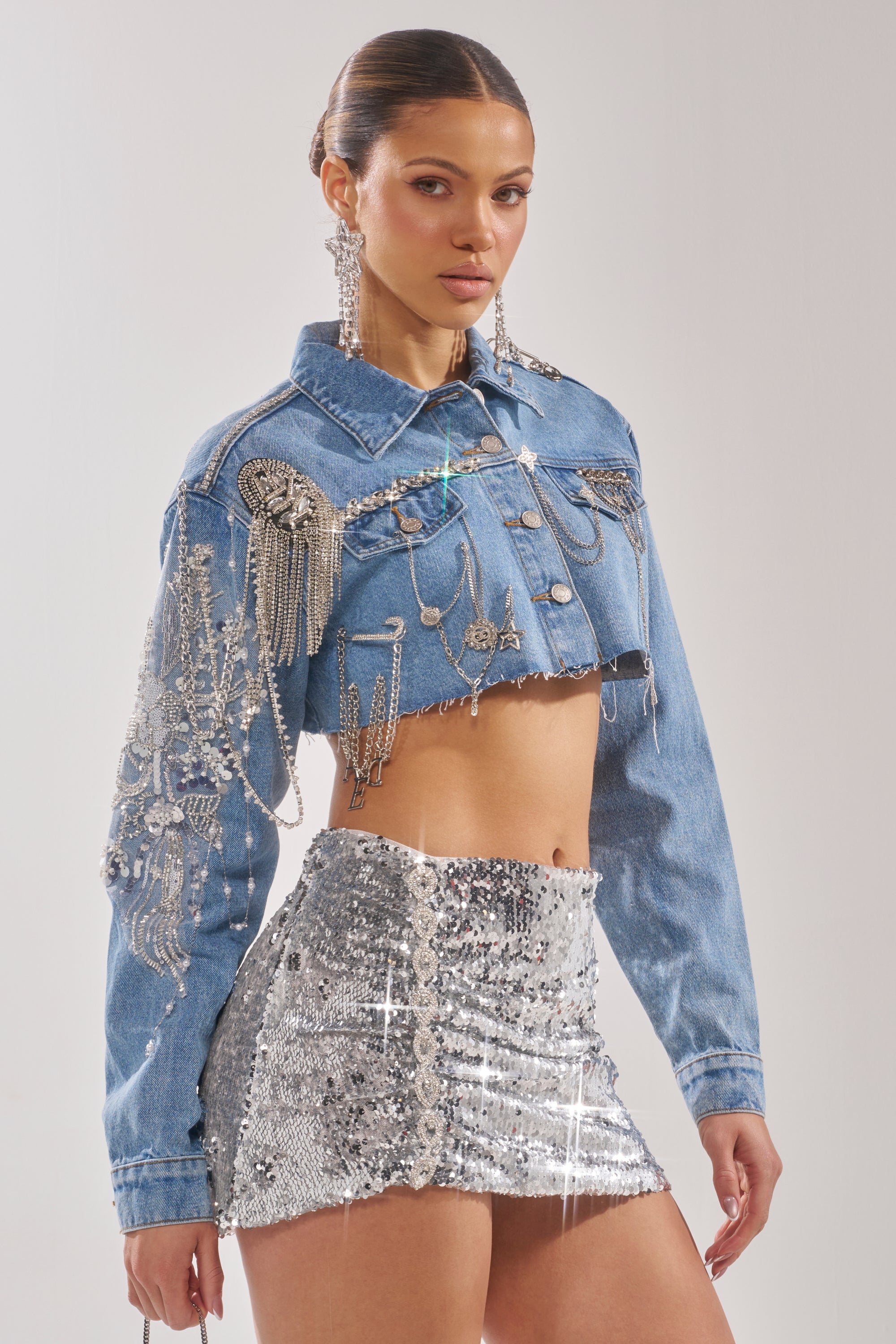 A woman poses confidently in the PIPER EMBELLISHED DENIM JACKET featuring silver fringe and jewels, styled with a sparkling silver sequin mini skirt and statement earrings against a plain background. 6