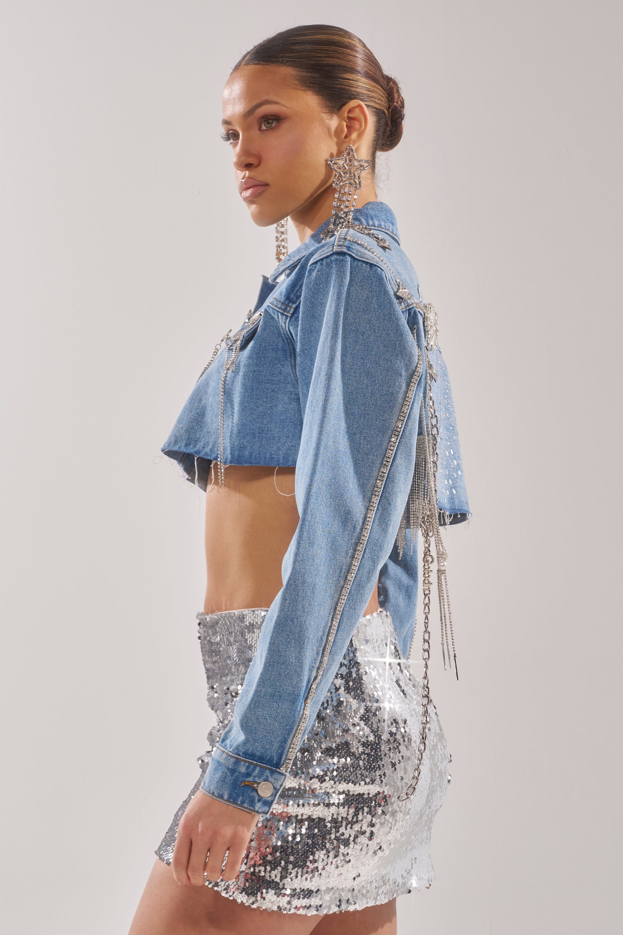 A woman poses confidently in the PIPER EMBELLISHED DENIM JACKET, featuring pearls and dangling chains, paired with sparkly earrings and a shiny silver sequin mini skirt. Her hair is in a sleek bun. 8