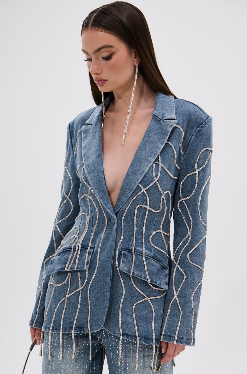 A woman models the STANDOUT EMBELLISHED DENIM BLAZER, featuring silver sequin wave patterns, large front pockets, and matching denim pants. She has long straight brown hair and statement earrings, looking down against a plain background.