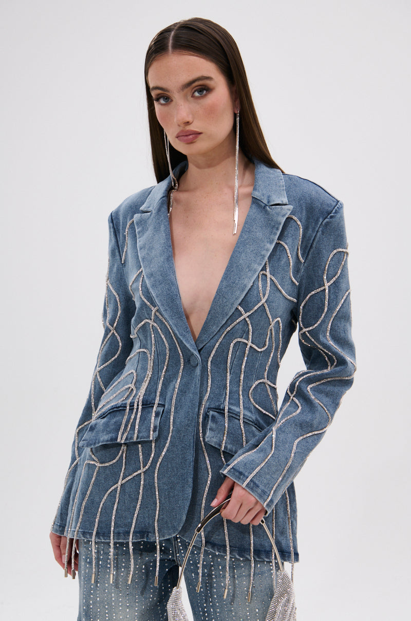 A woman with straight brown hair models the STANDOUT EMBELLISHED DENIM BLAZER, featuring wavy sequin details, worn without a shirt and paired with a small silver purse against a plain white background.