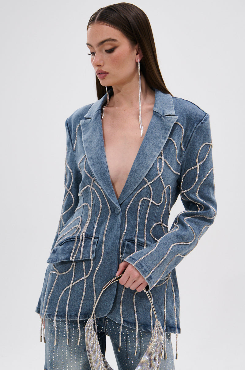 Wearing the STANDOUT EMBELLISHED DENIM BLAZER with matching jeans, long silver earrings, and a sparkling silver handbag, a woman stands against a plain light background, looking down.