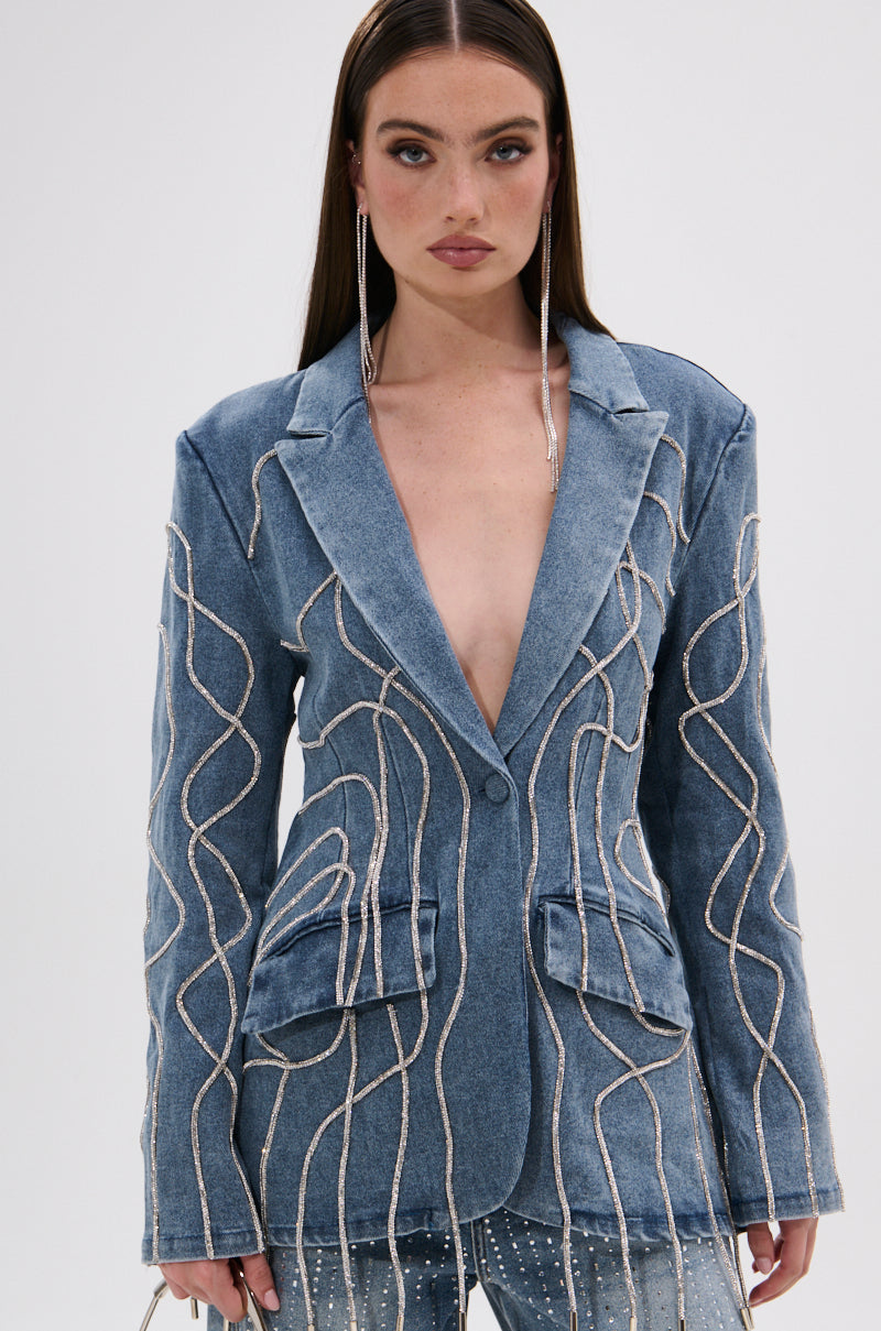 A woman with long brown hair wears the STANDOUT EMBELLISHED DENIM BLAZER, featuring wavy pearl and sequin details, over bare skin with matching rhinestone jeans, posing against a plain white background.