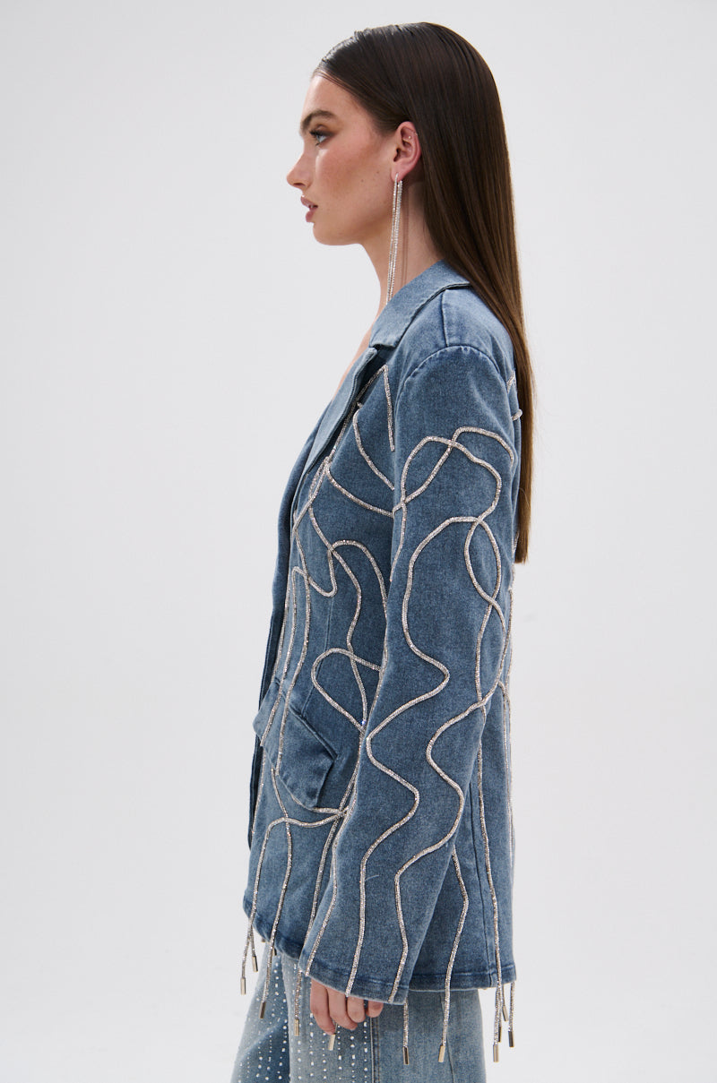 A woman with long, straight brown hair wears the STANDOUT EMBELLISHED DENIM BLAZER with wavy sequin details and matching denim pants. She faces sideways against a plain white background, accessorized with long earrings.