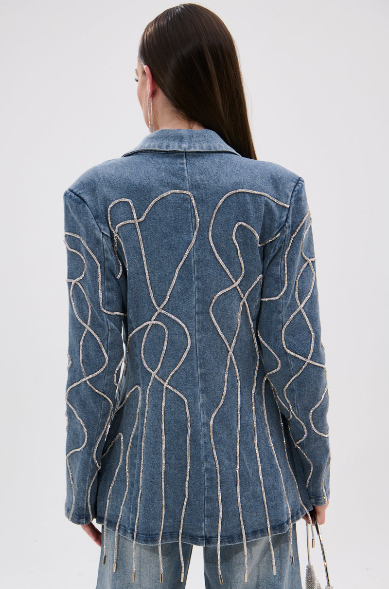 A person with long, straight brown hair models the STANDOUT EMBELLISHED DENIM BLAZER, featuring intricate beige embroidery and subtle sequin details on the back, against a plain white background.