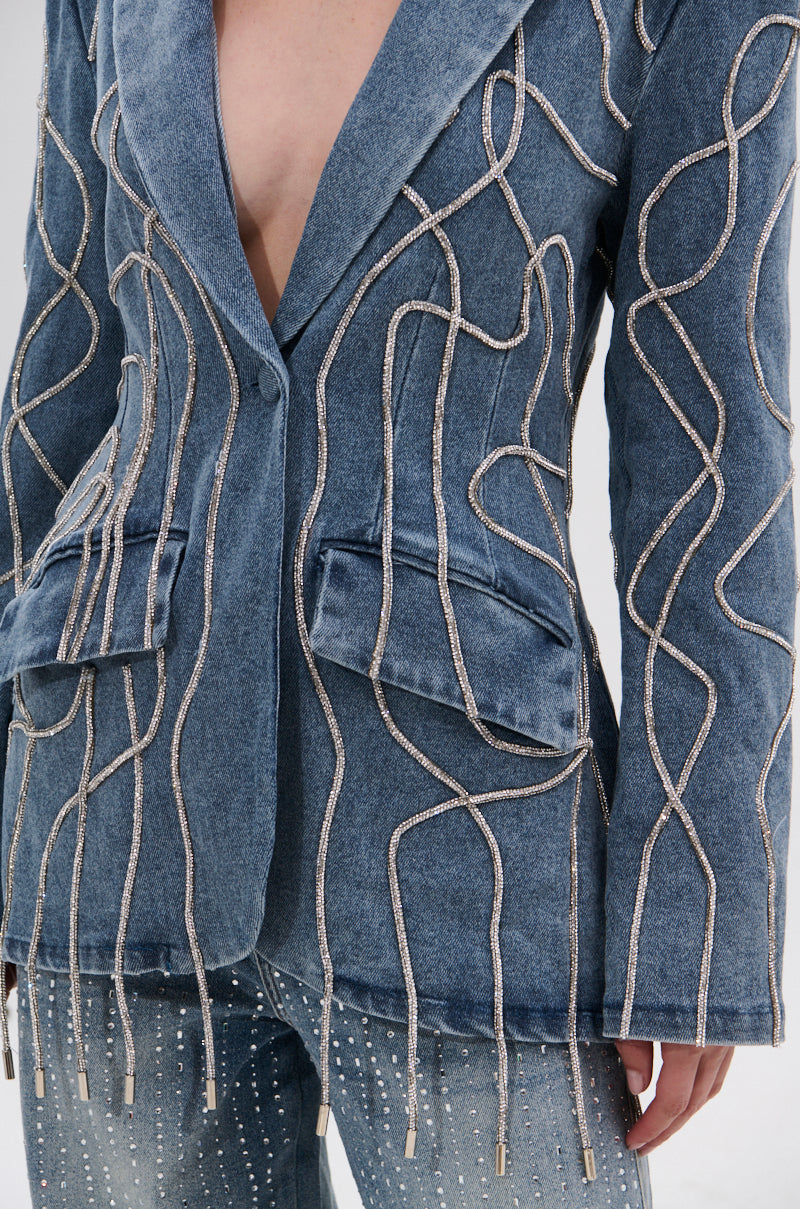 A person wears the STANDOUT EMBELLISHED DENIM BLAZER, a blue fitted piece with wavy sequin details, paired with matching studded denim pants and styled boldly buttoned without a shirt underneath.