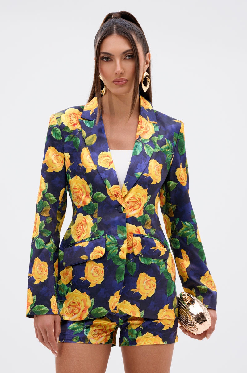 A woman styles the DOZEN ROSES SATIN BLAZER IN NAVY and matching shorts, featuring a bold yellow rose print. She pairs the look with large gold earrings, a gold textured clutch, and wears her long hair in a sleek high ponytail.