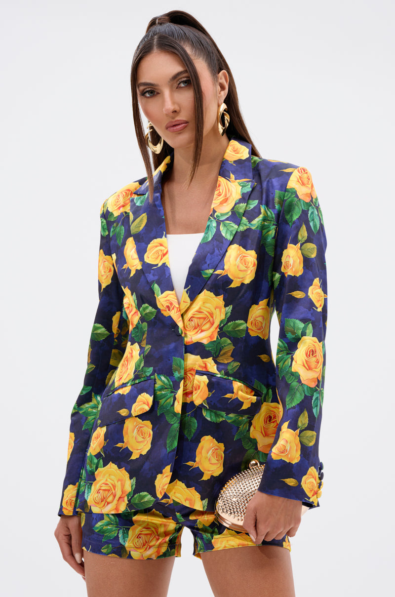 A woman wears the DOZEN ROSES SATIN BLAZER IN NAVY with matching shorts featuring a yellow rose print, paired with a white top, gold hoop earrings, and a gold clutch. Her long hair is in a high ponytail and she has a serious expression.
