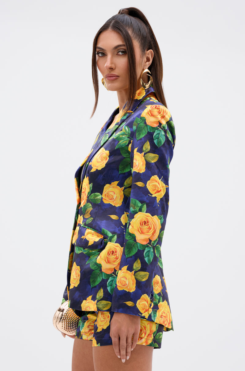 A woman with long dark hair in a high ponytail wears the DOZEN ROSES SATIN BLAZER IN NAVY with bold yellow rose print, matching shorts, gold hoop earrings, and holds a small gold clutch against a white background.