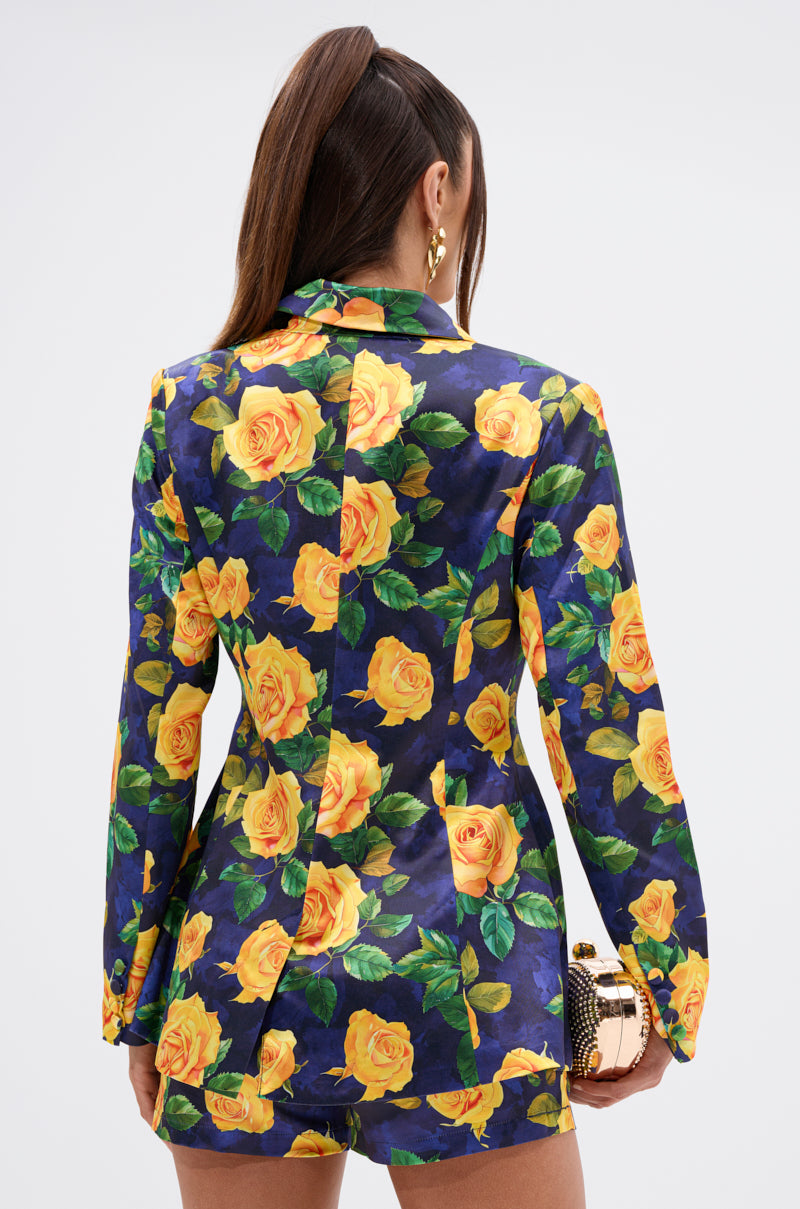 A woman with long brown hair in a ponytail wears the DOZEN ROSES SATIN BLAZER IN NAVY with bold yellow rose print, matching shorts, gold earrings, and a gold-and-black clutch. She faces away from the camera against a plain background.