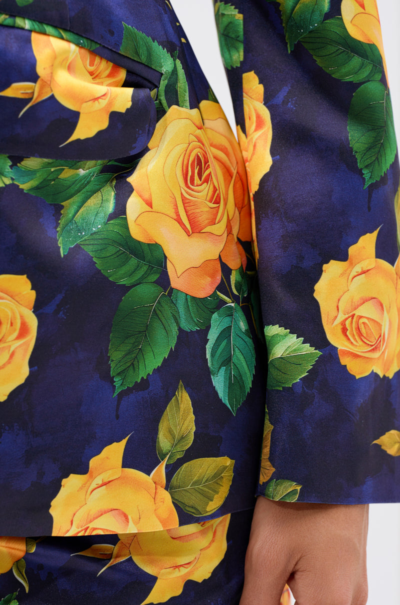 A close-up of a person wearing the DOZEN ROSES SATIN BLAZER IN NAVY, a dark blue blazer and pants set with a striking yellow rose print. The person's hand rests at their side, partially visible at the image's bottom.