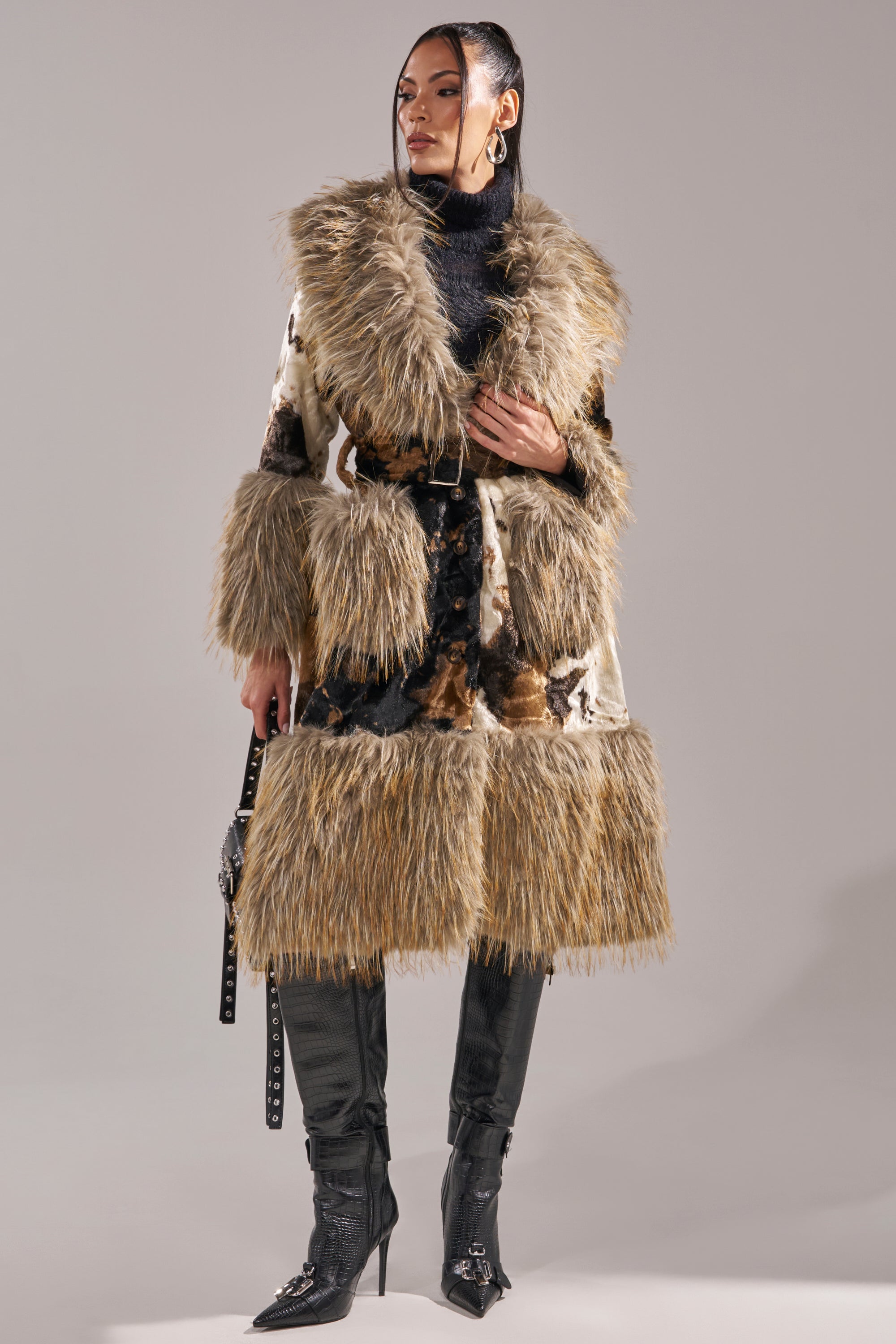 A woman wears the FREYA FUR TRIM TRENCH—a longline faux fur coat with wide collar and cuffs, belted at the waist—over a black turtleneck, black pants, and heeled boots, set against a plain gray background.