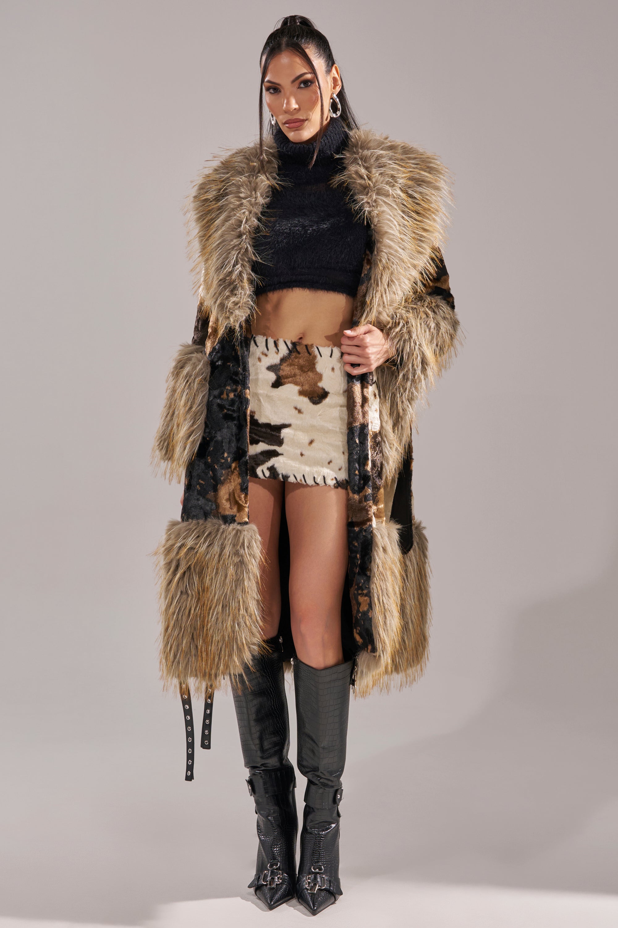 A woman stands confidently against a light gray background, wearing tall black boots, a black crop top, cowhide mini skirt, and the FREYA FUR TRIM TRENCH—a faux fur coat with brown and tan accents.