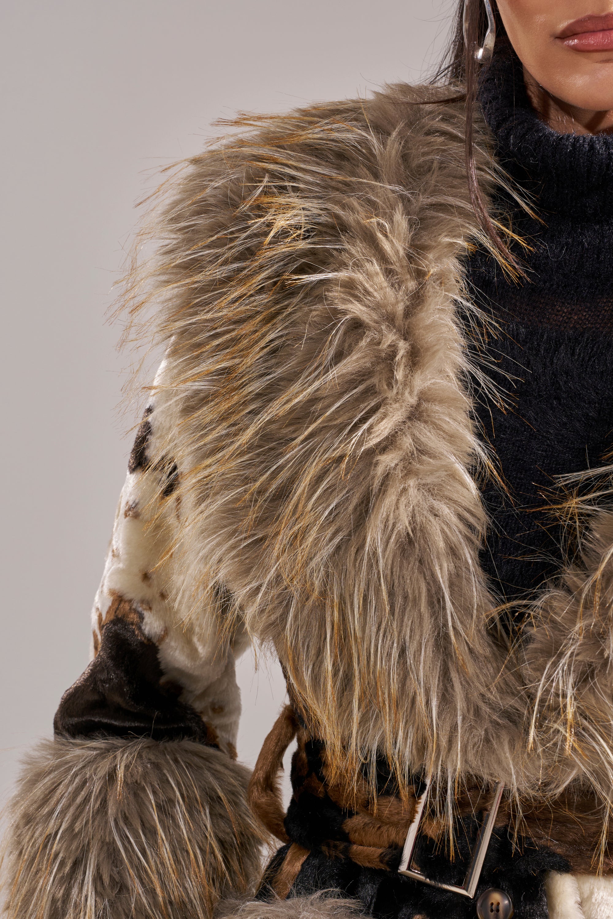 A close-up of a person wearing the FREYA FUR TRIM TRENCH, a shaggy faux fur coat in brown and beige over a black turtleneck, showing part of their face and torso.