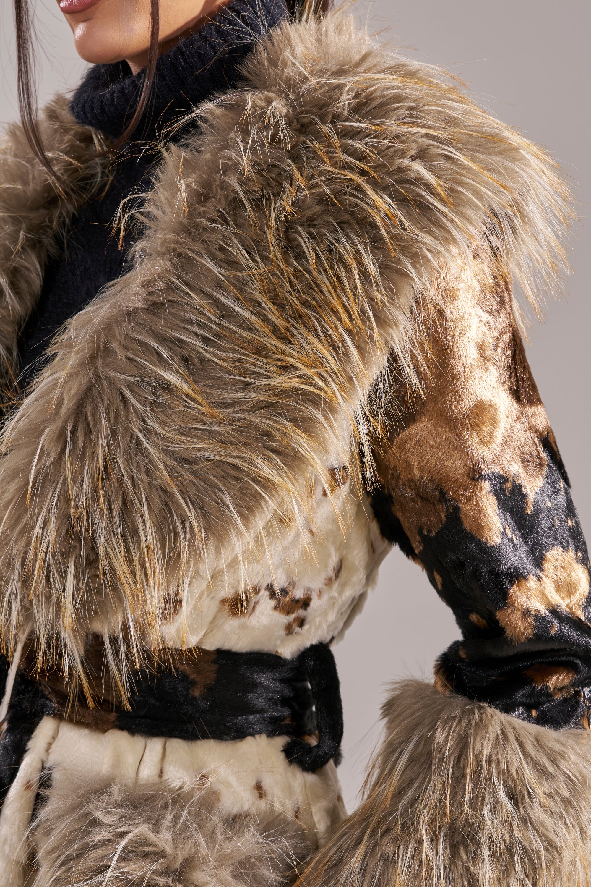 A person wearing the FREYA FUR TRIM TRENCH—cow print with a large, fluffy faux fur collar and cuffs, tied at the waist with a black belt—over a black turtleneck. Only the lower face and upper body are visible.