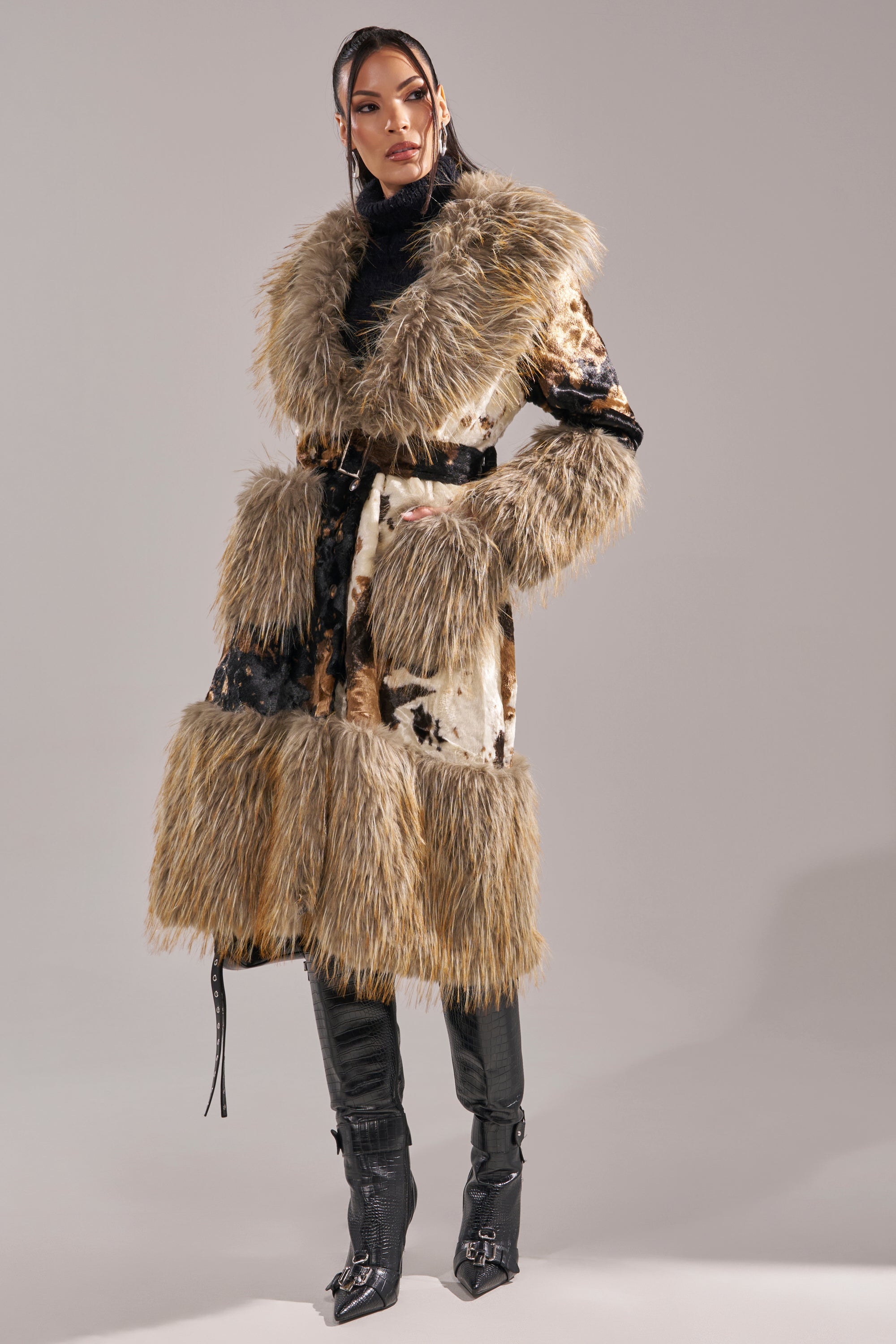 A person wearing the FREYA FUR TRIM TRENCH, styled with a black belt, turtleneck, and tall black boots, stands against a plain gray background.