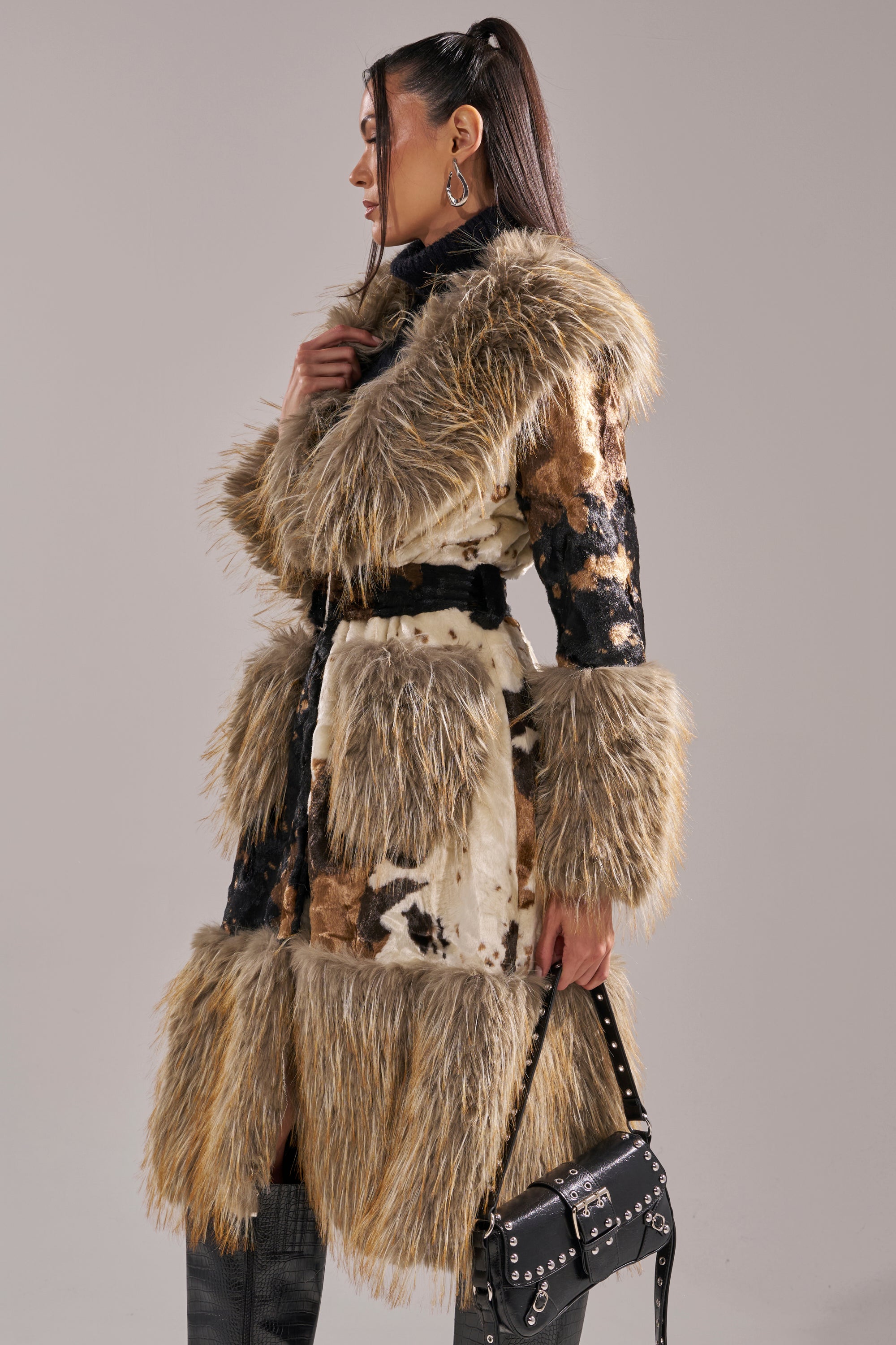 A woman in profile wears the FREYA FUR TRIM TRENCH, a cow print coat with fur trim, paired with hoop earrings and a black studded handbag. Her hair is styled in a high ponytail.