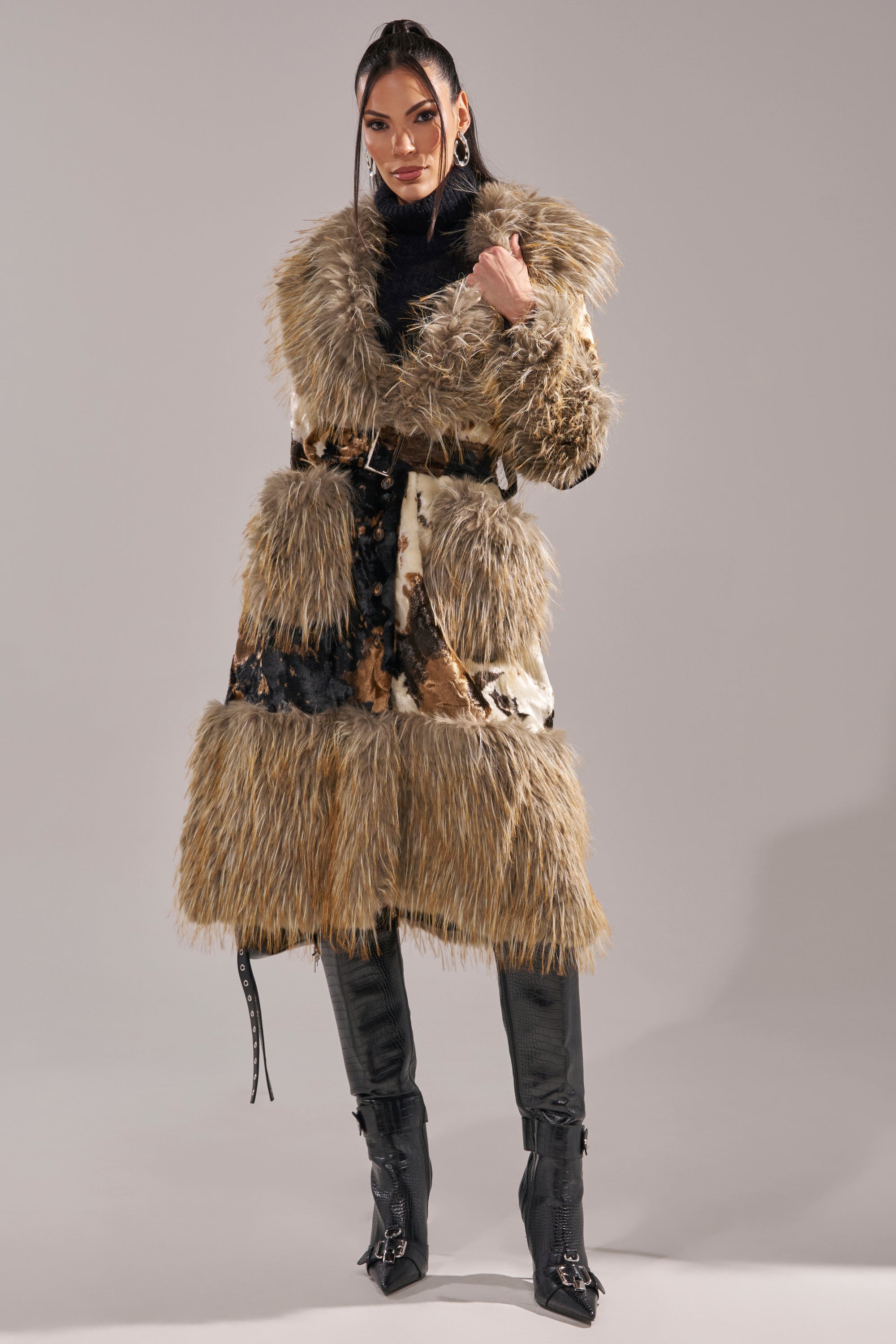 A woman stands confidently in the FREYA FUR TRIM TRENCH, styled with a black belt, black turtleneck, and knee-high black boots against a plain gray background.