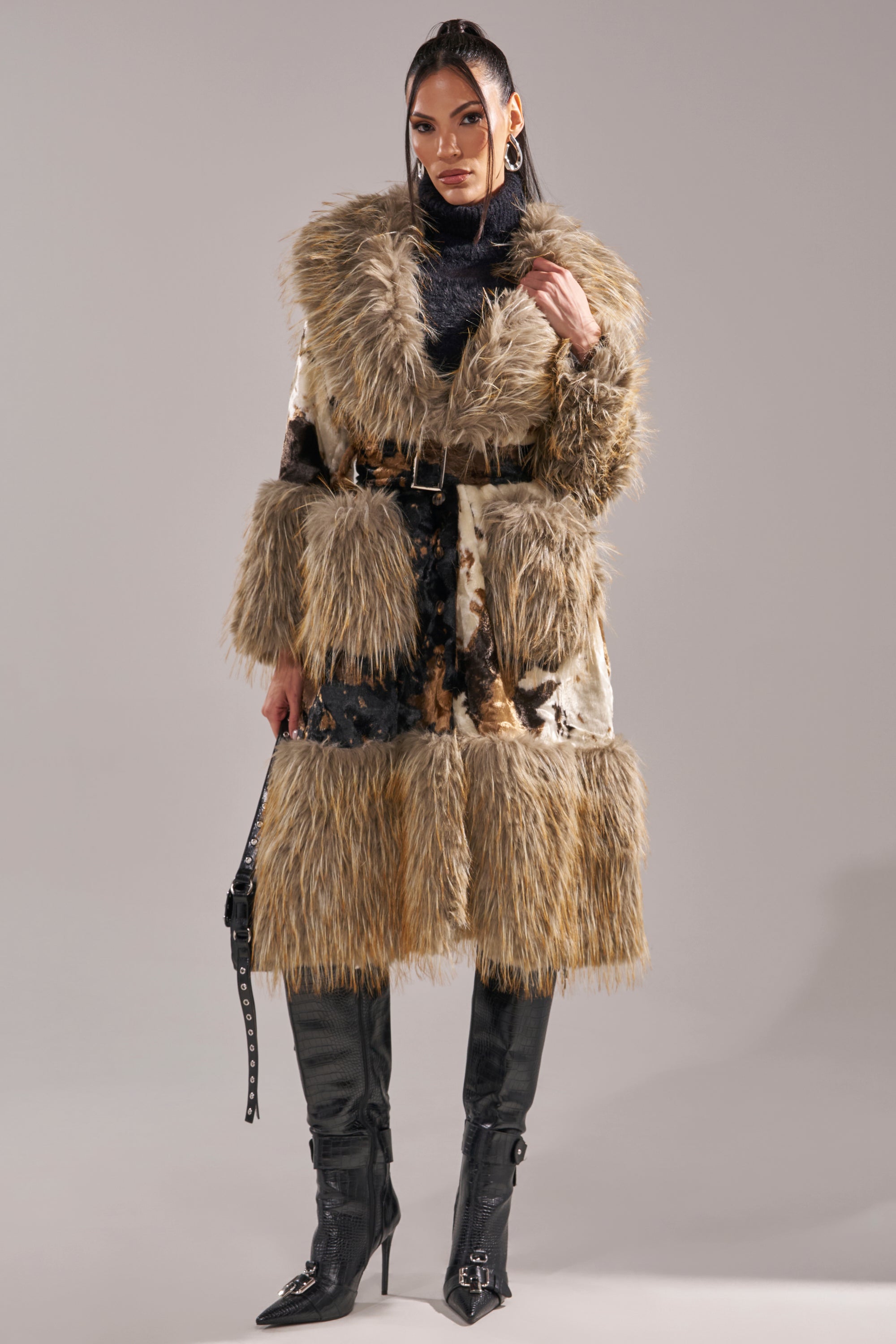 A person stands confidently in the FREYA FUR TRIM TRENCH, a longline faux fur coat in brown and beige tones, styled with a black turtleneck, fitted black boots, and a black handbag against a plain backdrop.