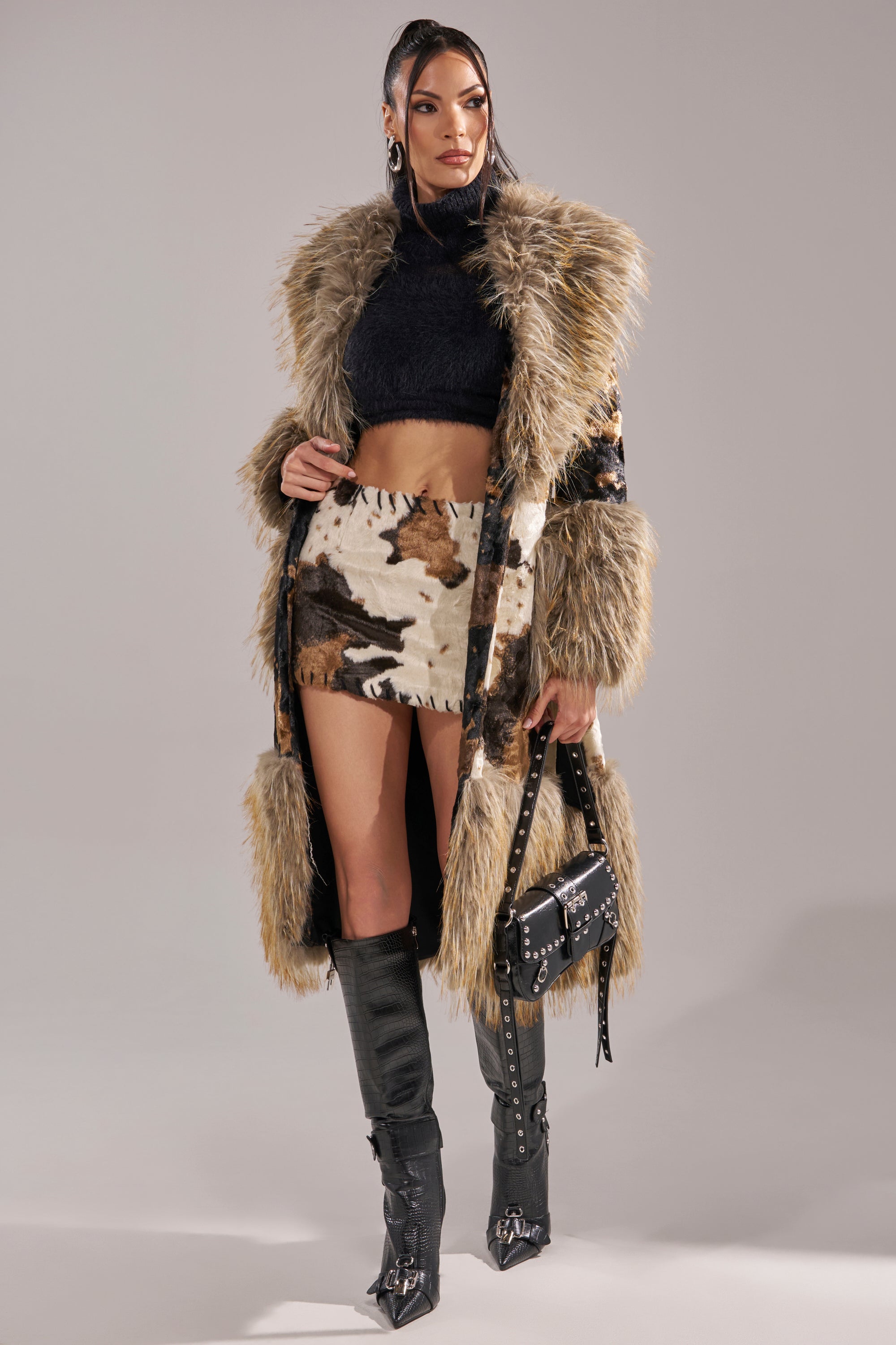 A woman stands confidently against a plain gray background, wearing the FREYA FUR TRIM TRENCH over a black crop top, cow print mini skirt, knee-high black boots, and holding a studded black handbag.