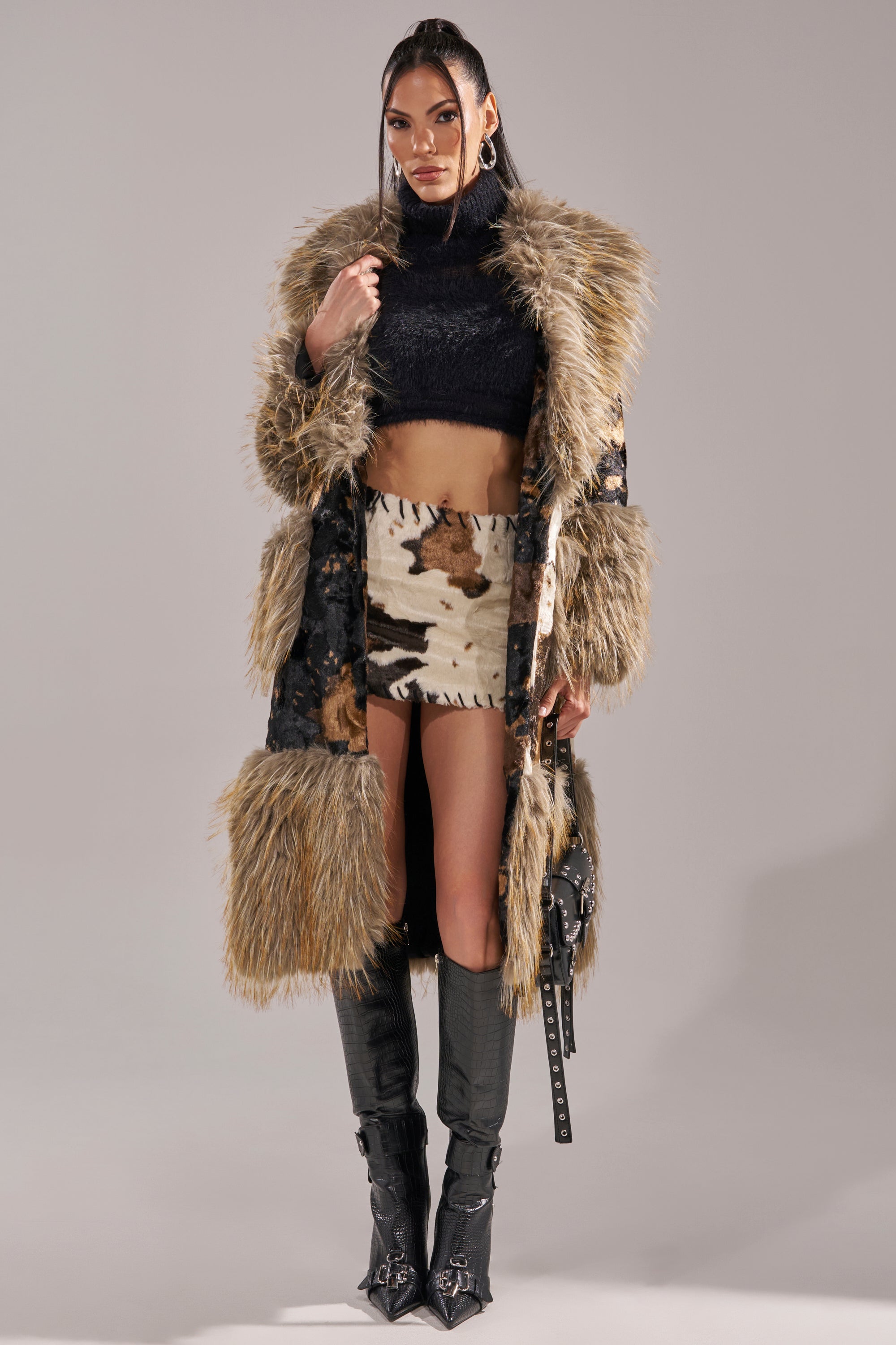 A woman wears the FREYA FUR TRIM TRENCH over a black crop top, cow-print mini skirt, and knee-high black boots. She carries a studded black handbag, her long straight hair styled with large hoop earrings.