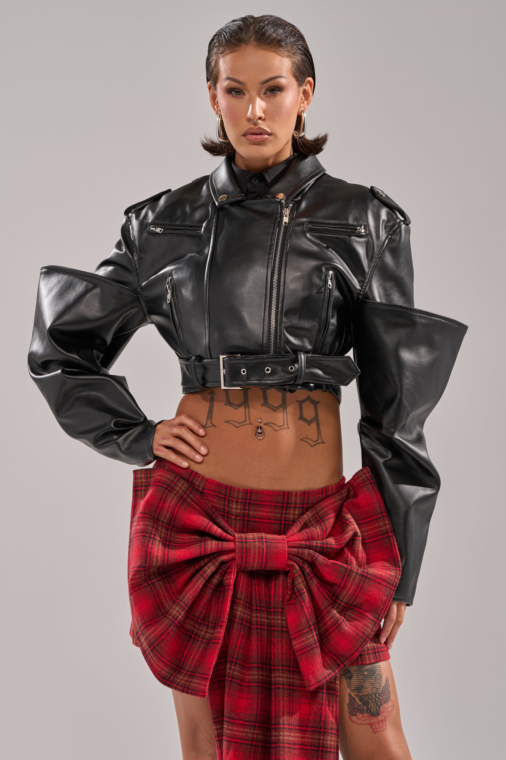A confident woman wears the STACK IT UP CROPPED MOTO in black faux leather, paired with a red plaid skirt featuring a large waist bow. Her slicked-back hair and visible stomach tattoos complete the edgy look. 0