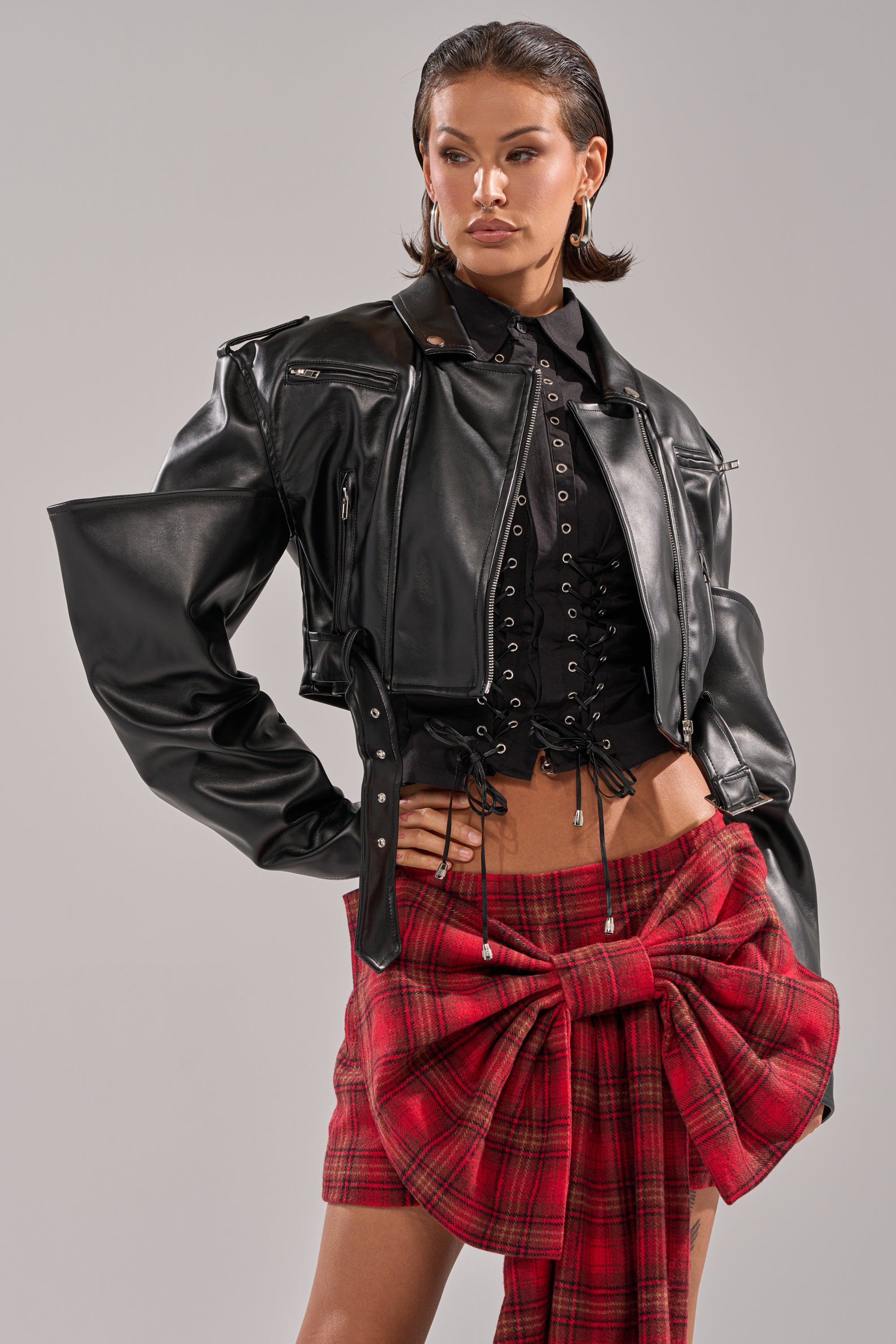 A woman wears the STACK IT UP CROPPED MOTO jacket over a black lace-up corset top and a red plaid skirt with an oversized bow, accessorized with gold hoop earrings, her hair slicked back as she poses confidently against a plain background. 1