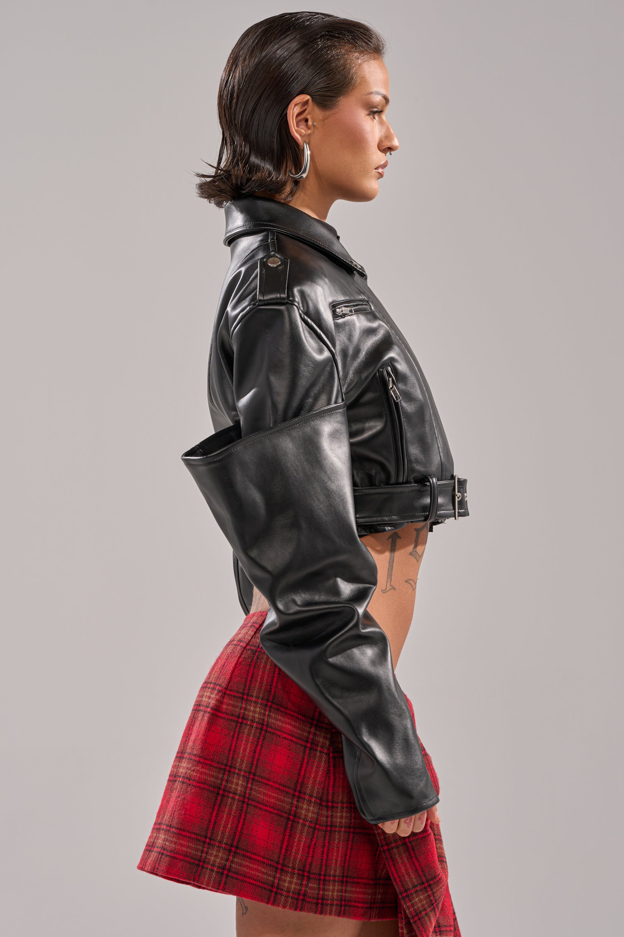 A woman with short brown hair stands in profile against a gray background, wearing the STACK IT UP CROPPED MOTO jacket with stacked sleeves draped off one shoulder and paired with a red plaid skirt. 4