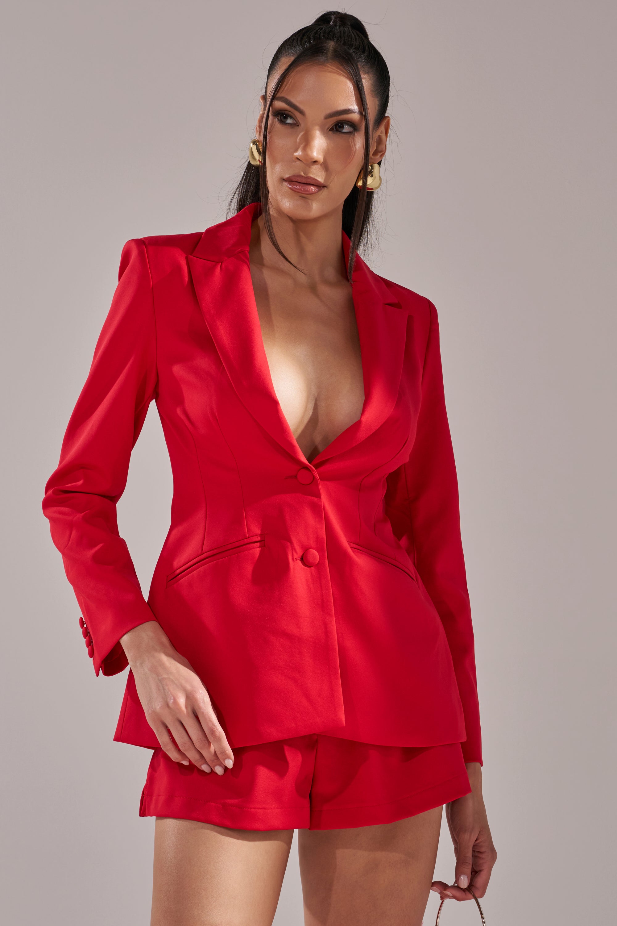 A woman exudes confidence in the CHERRY BOMB BLAZER, a bold women's blazer with a deep neckline paired with matching red shorts, her long dark hair swept back, gold hoop earrings on, and sunglasses in hand against a simple background. 2