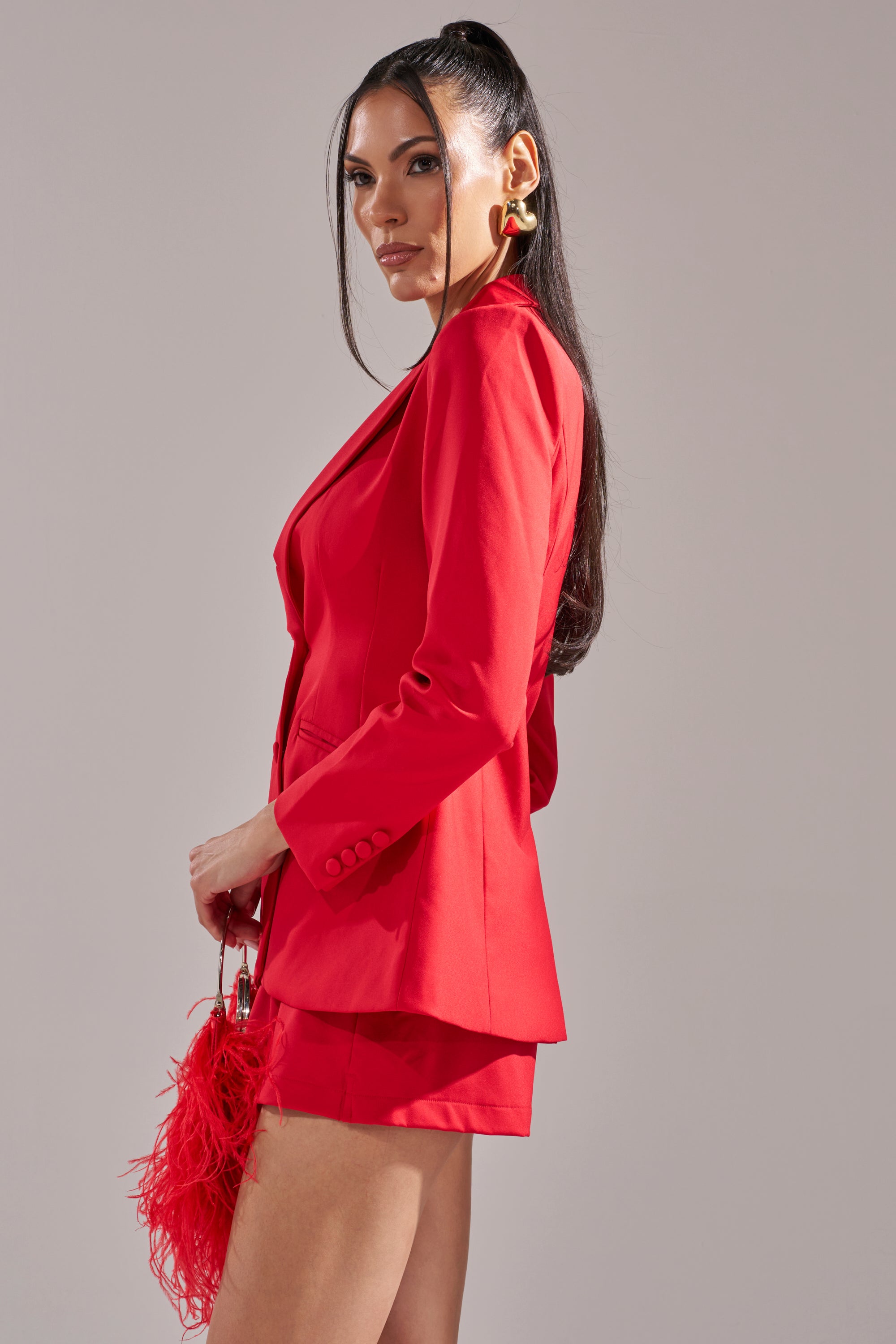 A woman with long dark hair in a high ponytail wears the CHERRY BOMB BLAZER and matching shorts, paired with a red feathered handbag, standing against a light gray background. 3