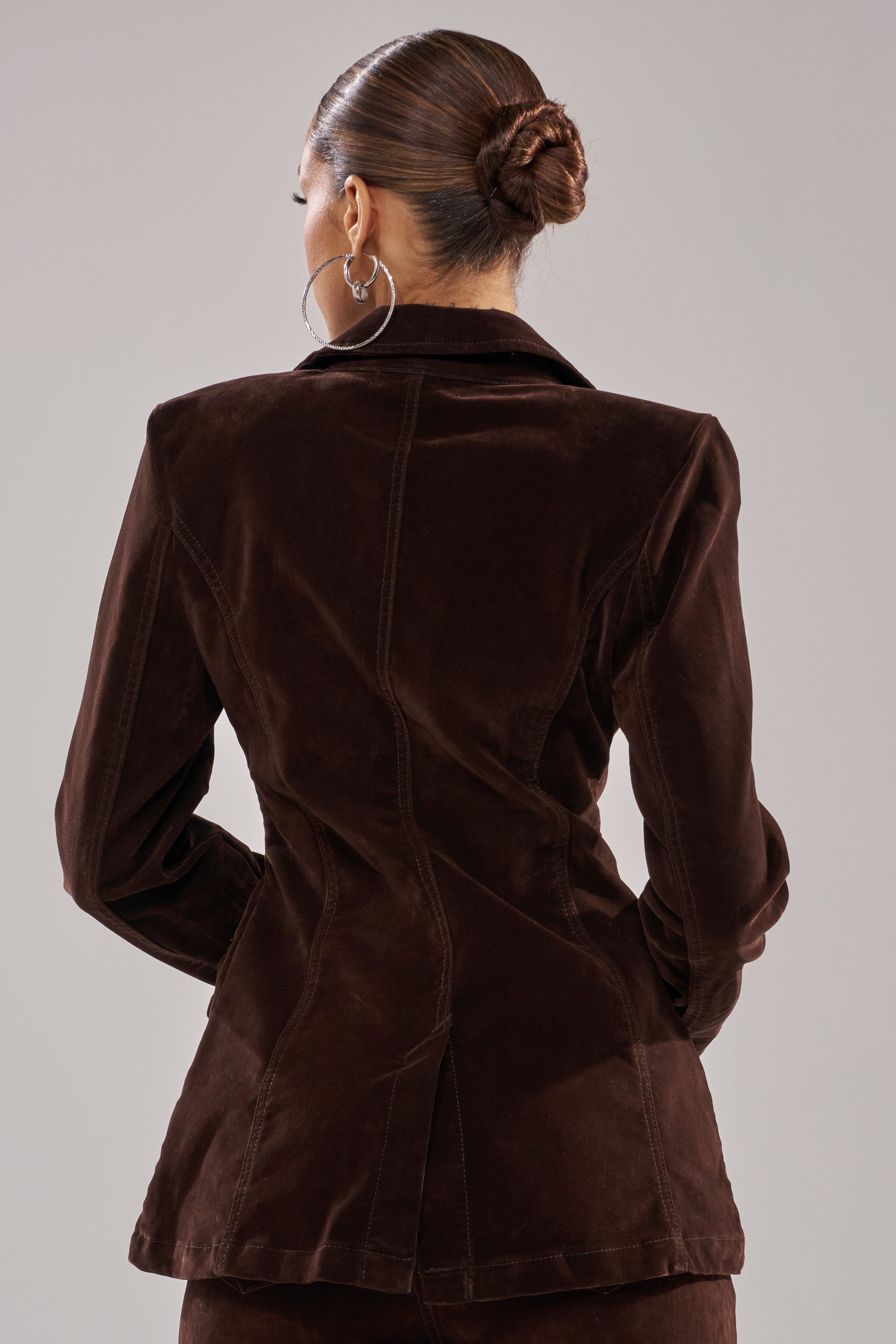 A person with their hair in a bun and large hoop earrings models the RAE VELVET DENIM BLAZER in dark brown, standing with their back to the camera against a neutral background. 3