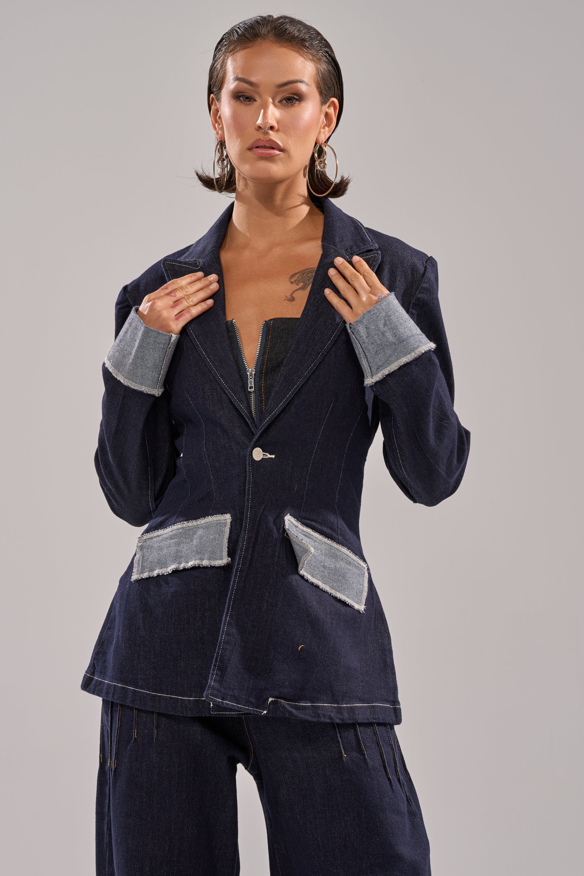 A person with slicked-back hair wears the JESSE DENIM BLAZER—dark denim with frayed light cuffs and pockets—styled with matching pants, posing confidently against a plain neutral background. 0
