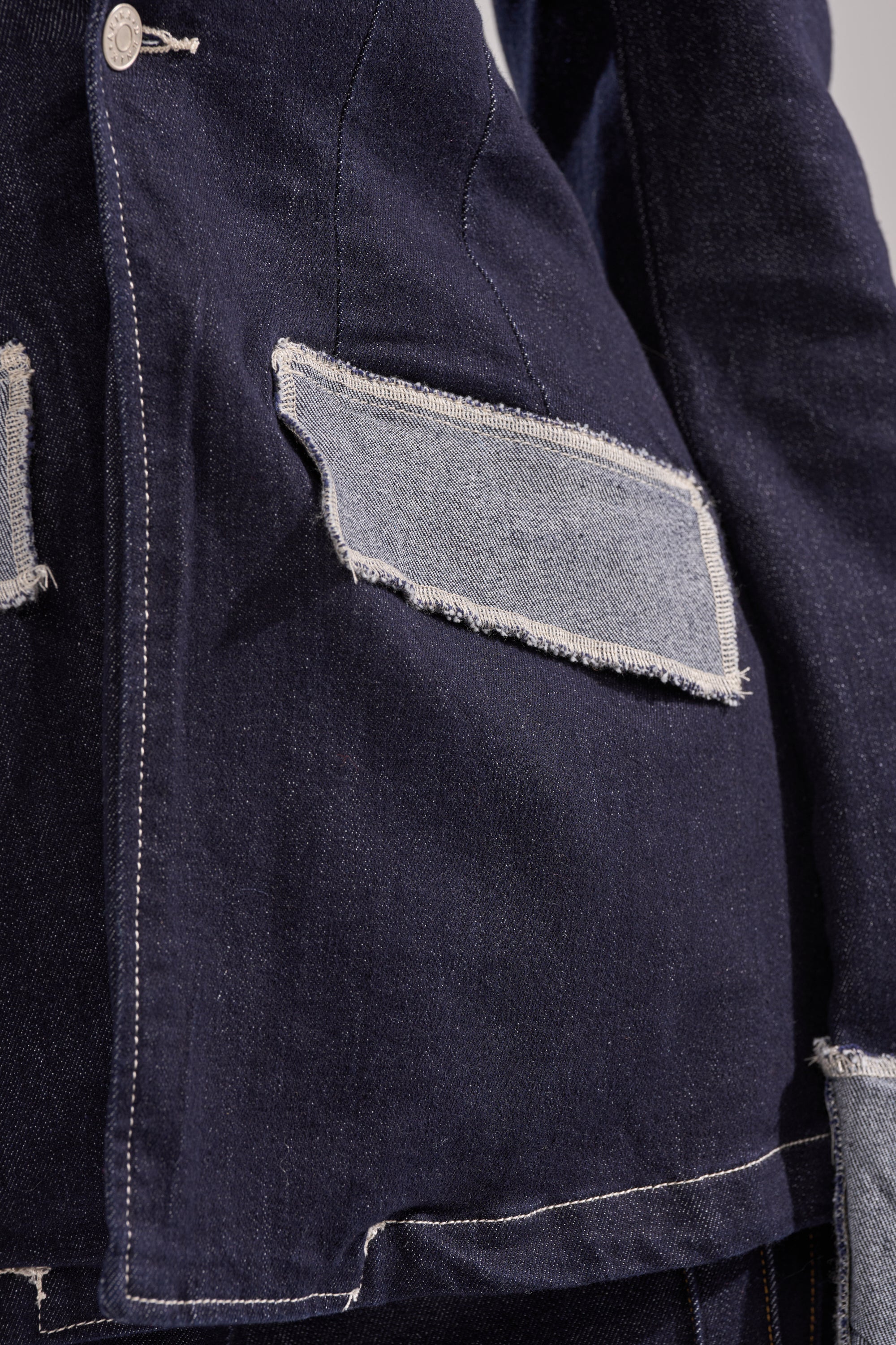 Close-up of the JESSE DENIM BLAZER—a dark denim jacket with modern tailoring, large raw-edge light denim patches on the pockets, contrast stitching, and metallic buttons for a structured silhouette. 9