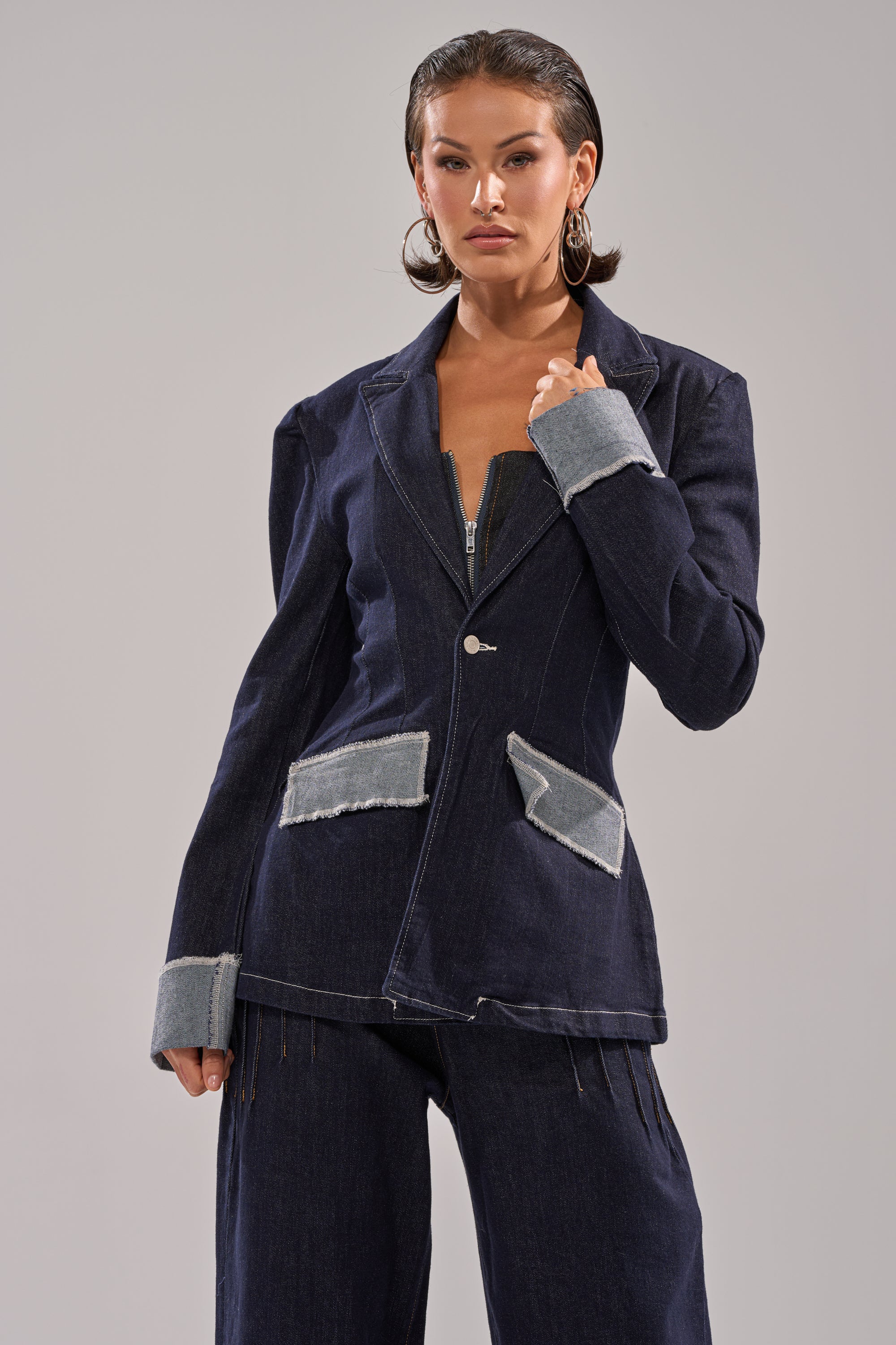 A woman with slicked-back hair wears the JESSE DENIM BLAZER, featuring a dark wash with light denim cuffs and pocket flaps. The fitted cut and frayed detailing create a structured look as she poses against a plain, light gray background. 1