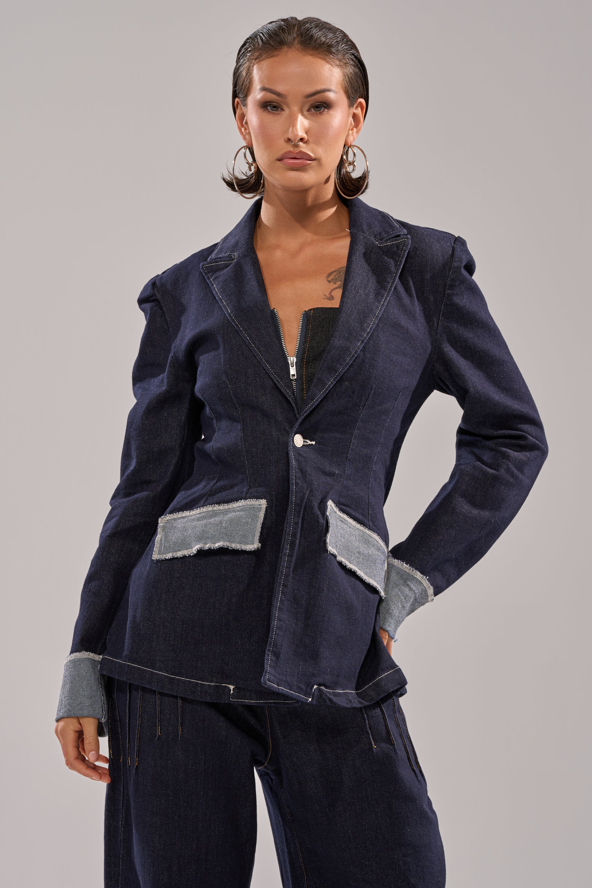 A woman with wet-look hair wears the JESSE DENIM BLAZER, showcasing modern tailoring and light denim details, paired with matching pants as she stands confidently against a plain gray background. 2