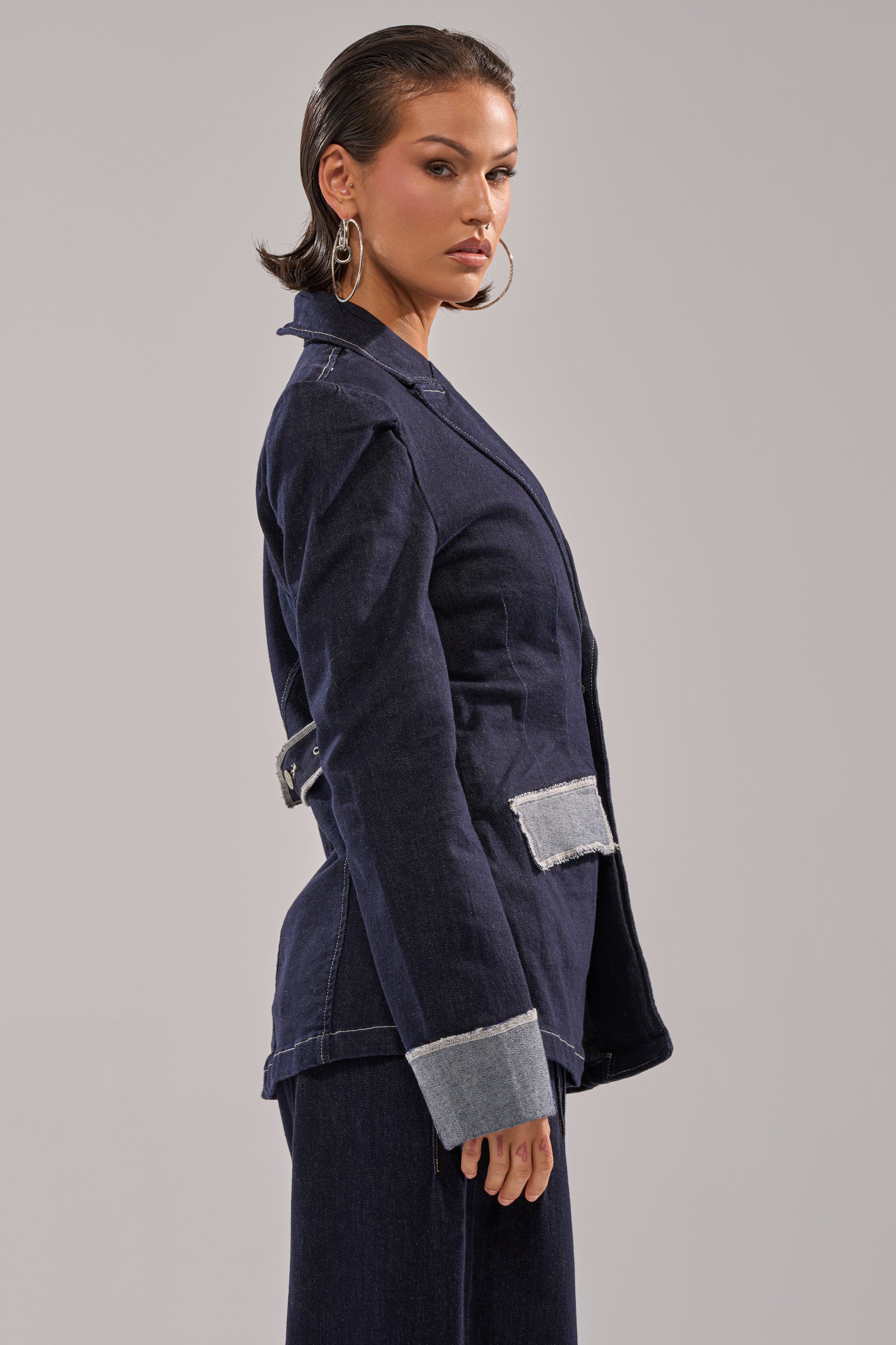 Woman in profile against a light background wears the JESSE DENIM BLAZER, featuring dark denim with light denim cuffs and pocket flaps, styled with large hoop earrings and slicked-back hair to highlight its modern, structured tailoring. 3