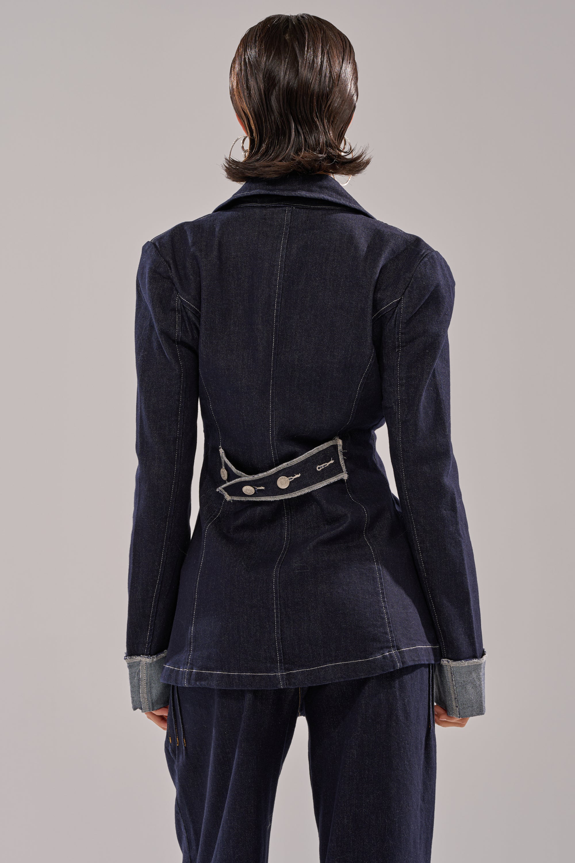 A person with short, slicked-back hair is seen from behind wearing the JESSE DENIM BLAZER with an adjustable strap and matching pants, featuring rolled-up sleeves that reveal a contrasting lighter denim lining. 4
