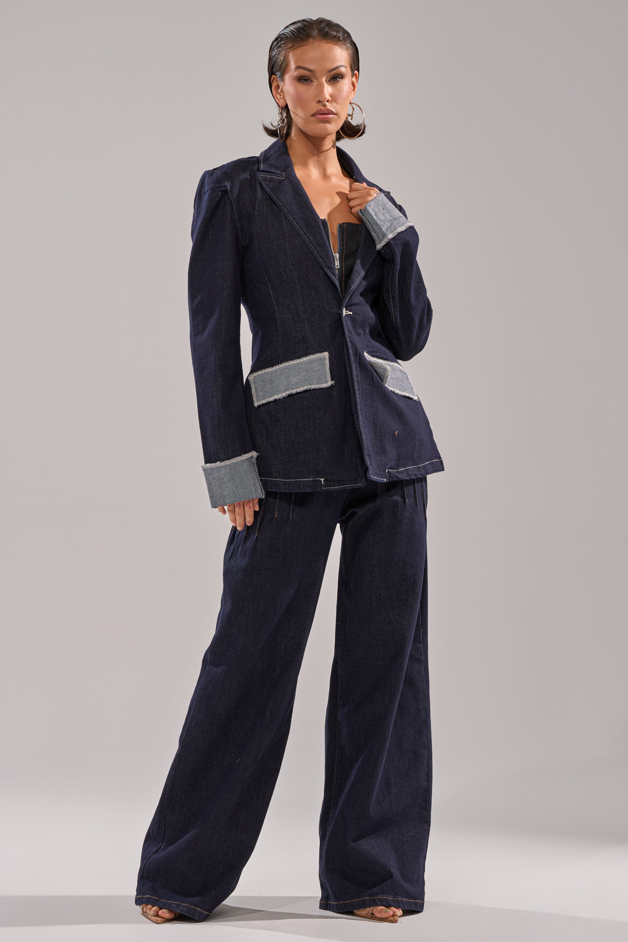 A fashion-forward individual poses confidently in the JESSE DENIM BLAZER—a structured dark denim pantsuit with light grey cuff and pocket accents, styled with large hoop earrings and slicked-back hair against a plain background. 5