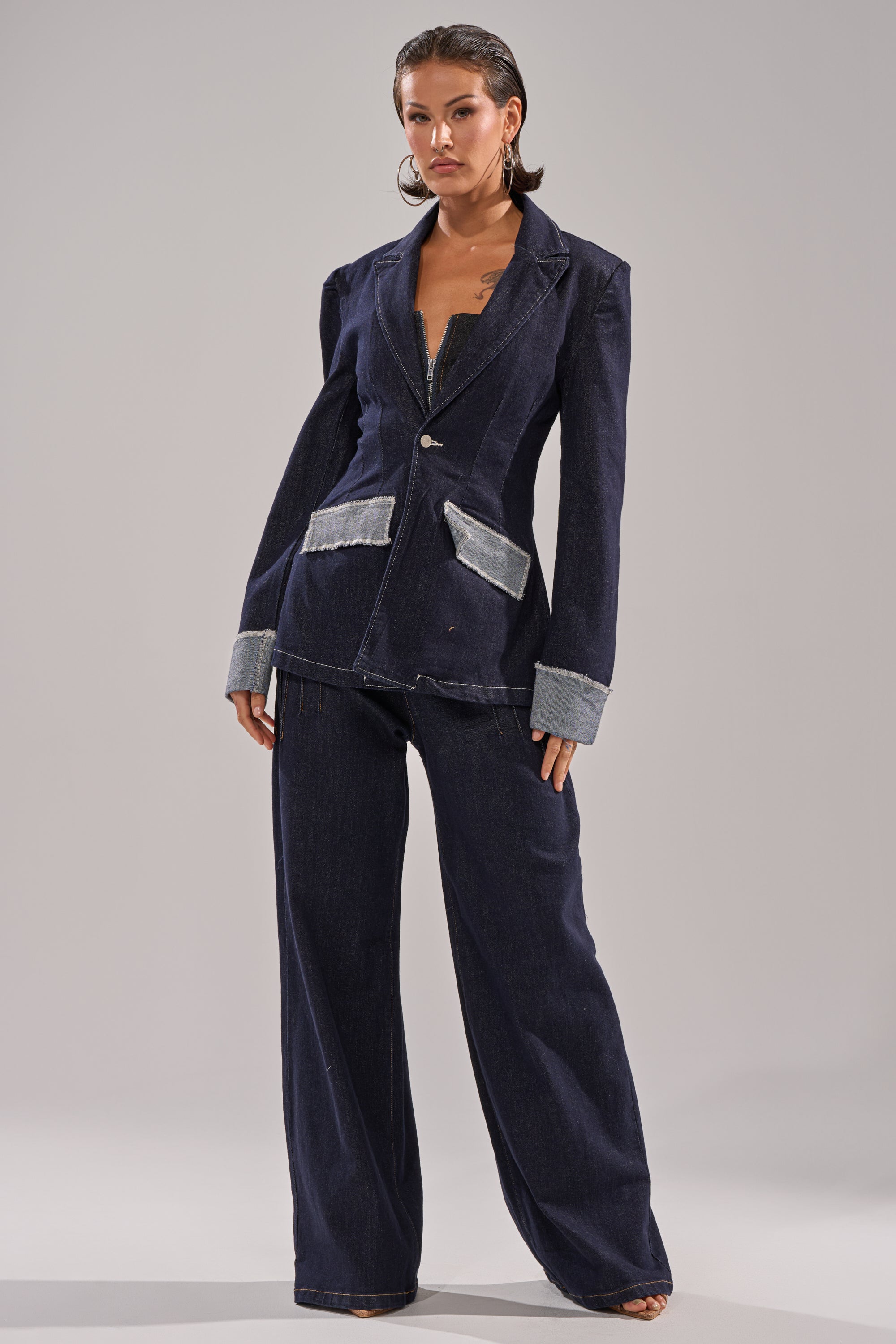 A woman stands confidently in the JESSE DENIM BLAZER, styled with wide-leg pants and featuring light denim accents on the cuffs and pockets, showcasing modern tailoring against a plain light gray background. 6