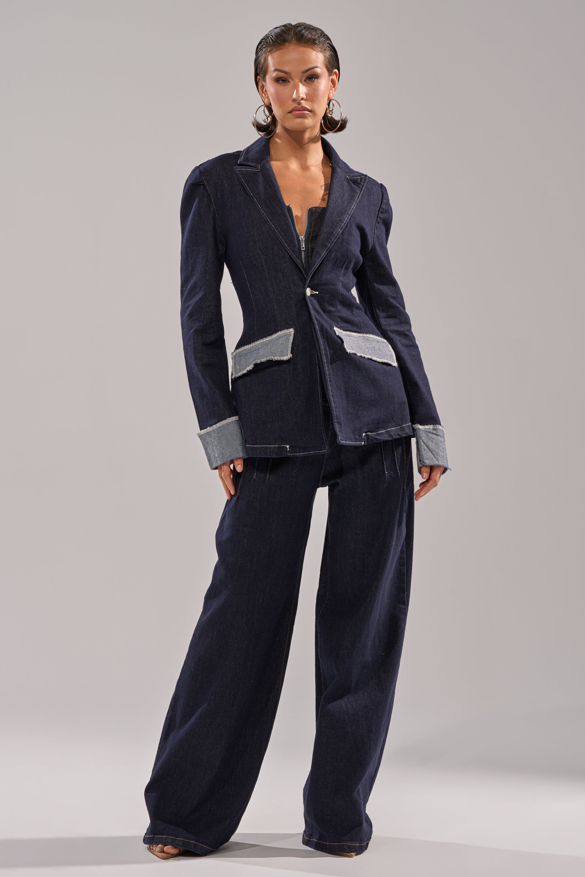 A woman stands confidently in modern tailoring, wearing the JESSE DENIM BLAZER with light denim accents and wide-leg pants, creating a structured silhouette as she poses against a plain gray background. 8
