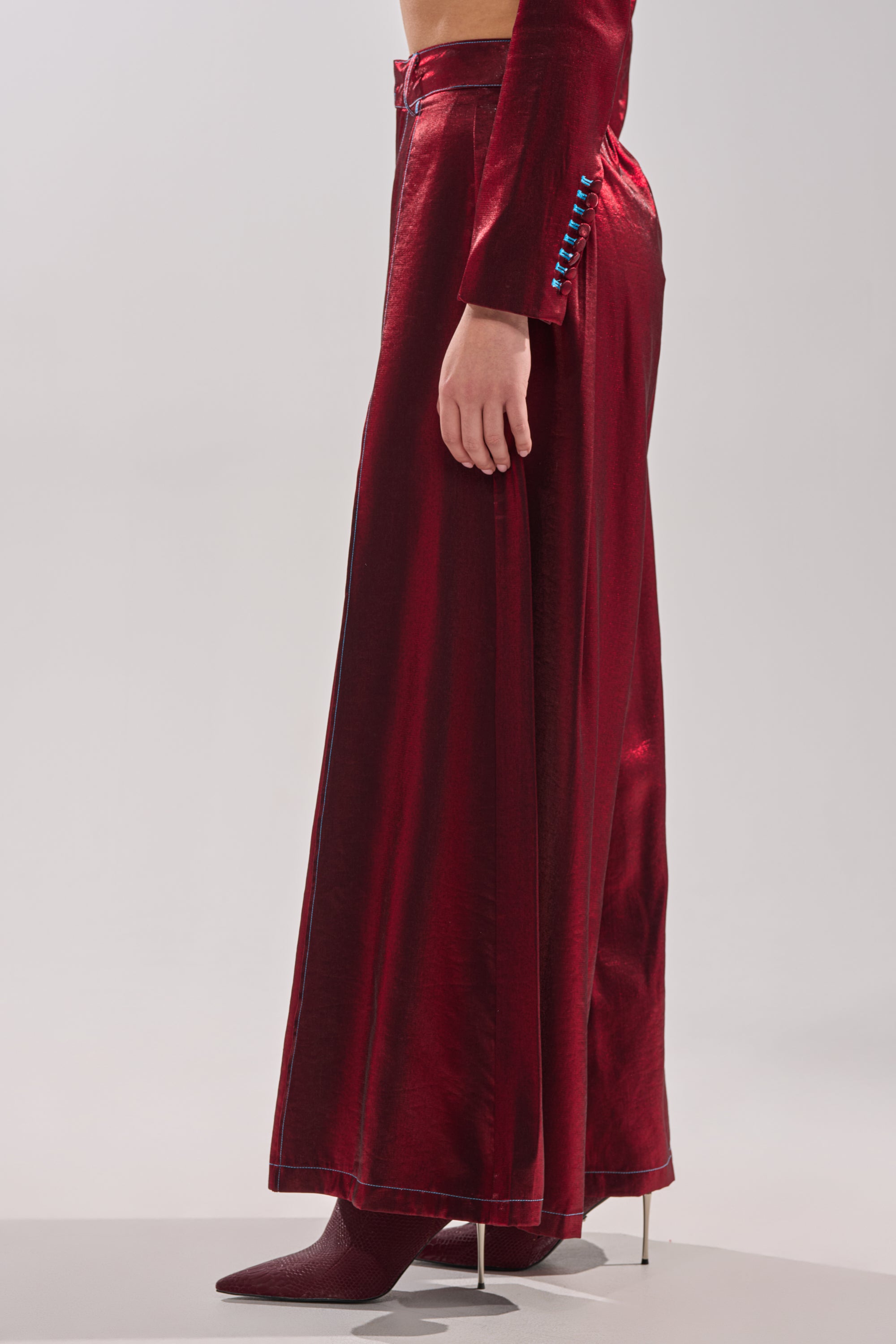 Someone wears the MISS YOU MOST WIDE LEG PANT in satin burgundy, styled with matching pointed-toe heels. Blue buttons run along the side seam, all set against a plain, light background. 1