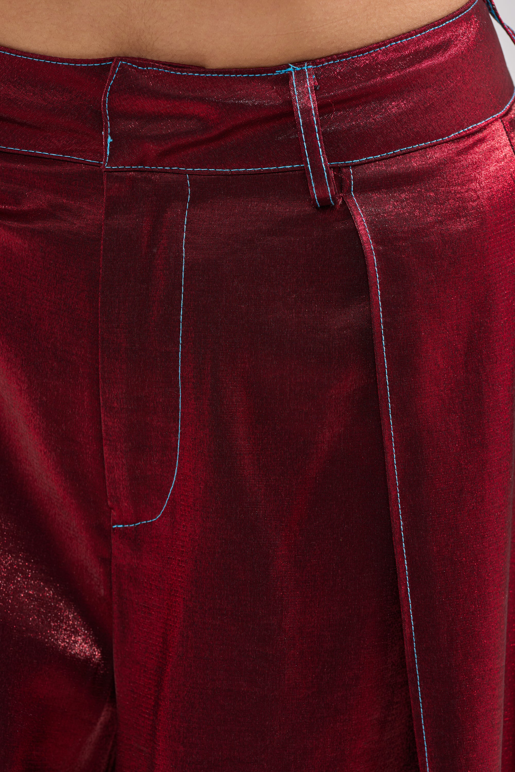 Close-up of a person wearing the MISS YOU MOST WIDE LEG PANT—high-waisted, metallic red satin with bold blue stitching at the waistband, fly, and pockets. The glossy burgundy fabric catches the light for a striking look. 4