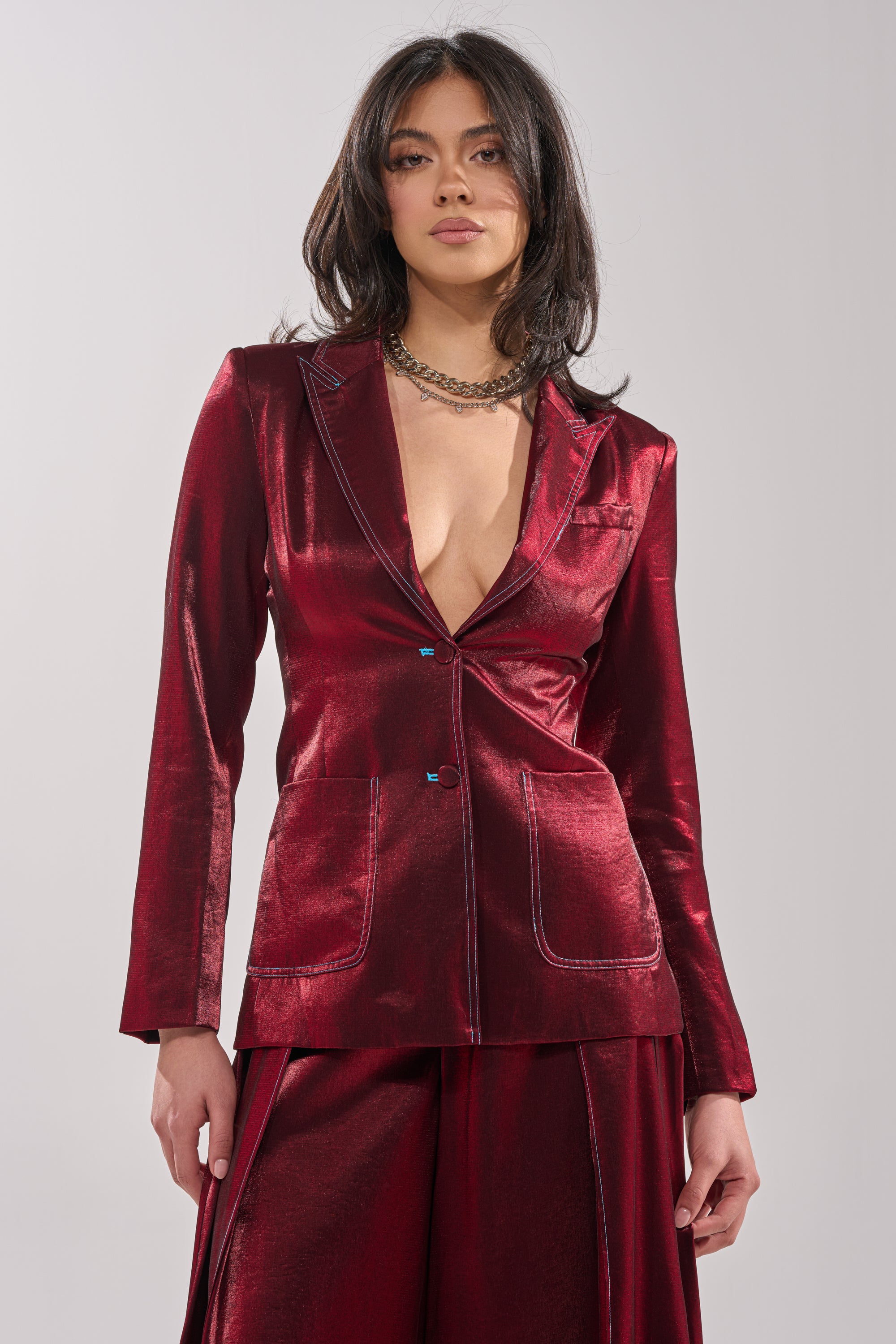 A woman radiates confidence in the MISS YOU MOST BLAZER—a deep red, satin-finish piece with wide lapels and front pockets—styled with matching bottoms, loose dark hair, and a chunky chain necklace. 1